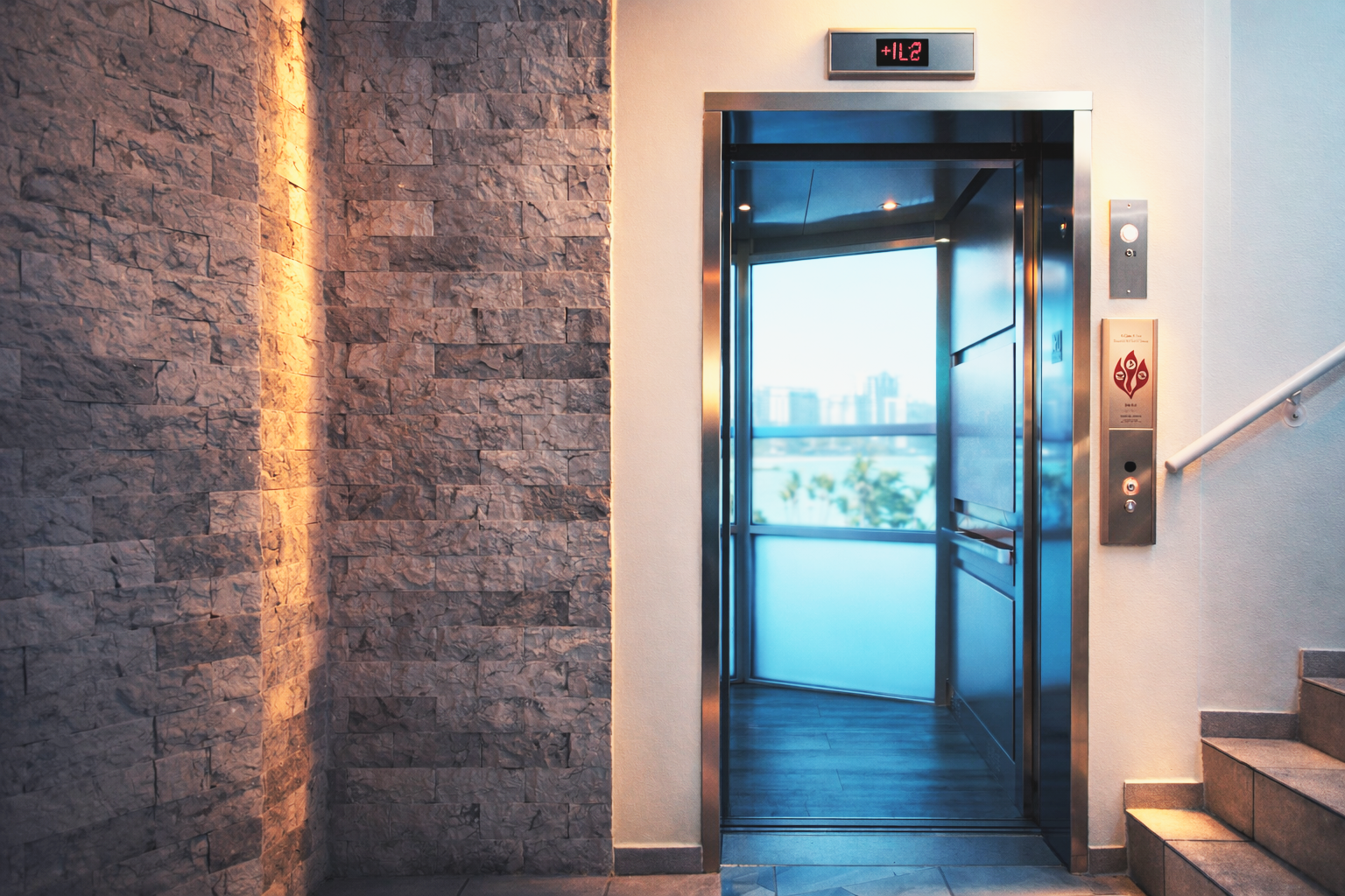 Ascensor abierto en un edificio con vista a la ciudad y un reloj digital que muestra +11:22.
