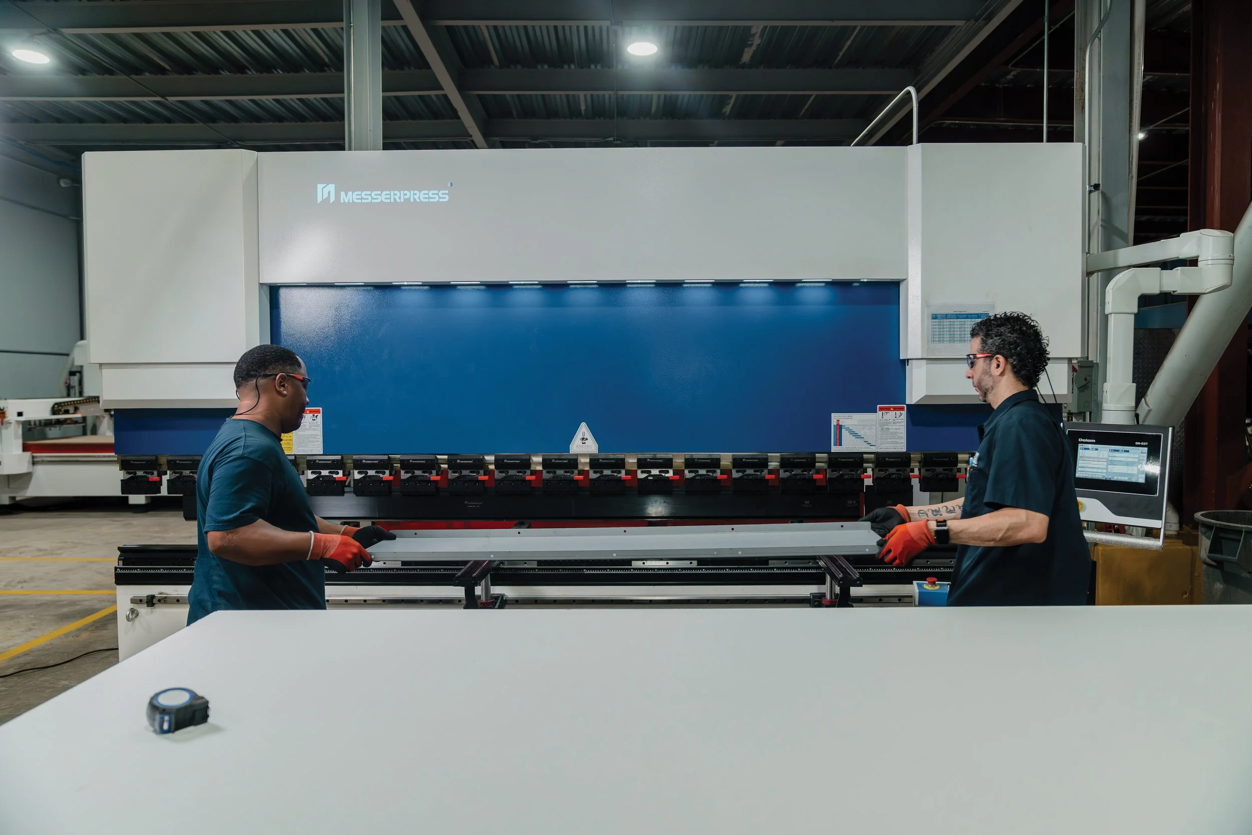 Dos trabajadores en una fábrica operando una máquina grande de fabricación de metal, con el logo de Messerpress en la máquina.