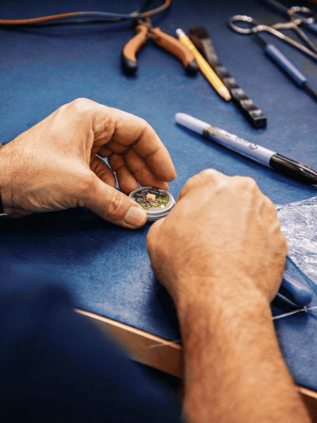 Manos manipulando un microchip o circuito en una superficie azul, con varias herramientas y componentes electrónicos alrededor.