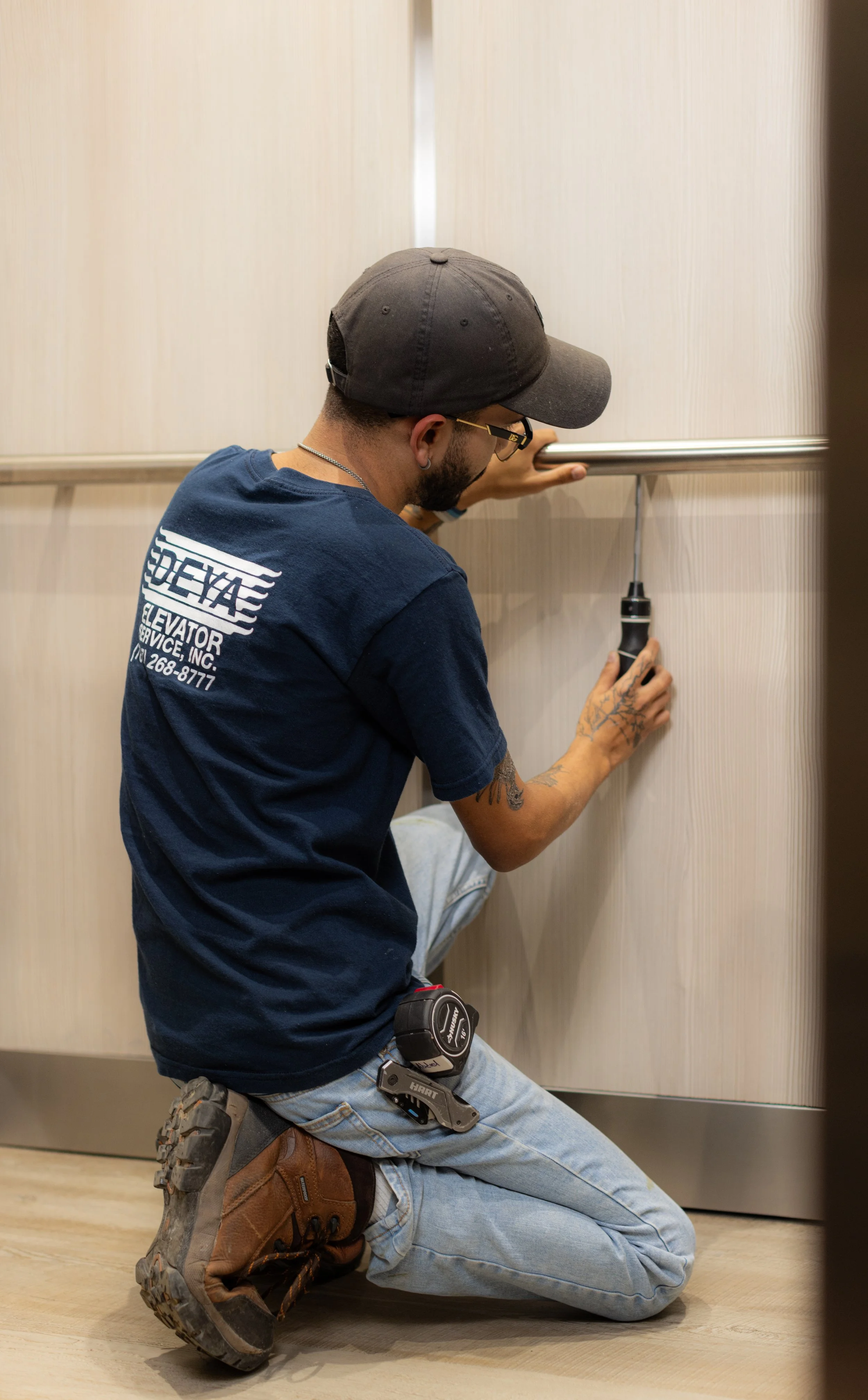 Un trabajador en posición de cuclillas, usando un martillo para ajustar o reparar una estructura en una pared de un ascensor.