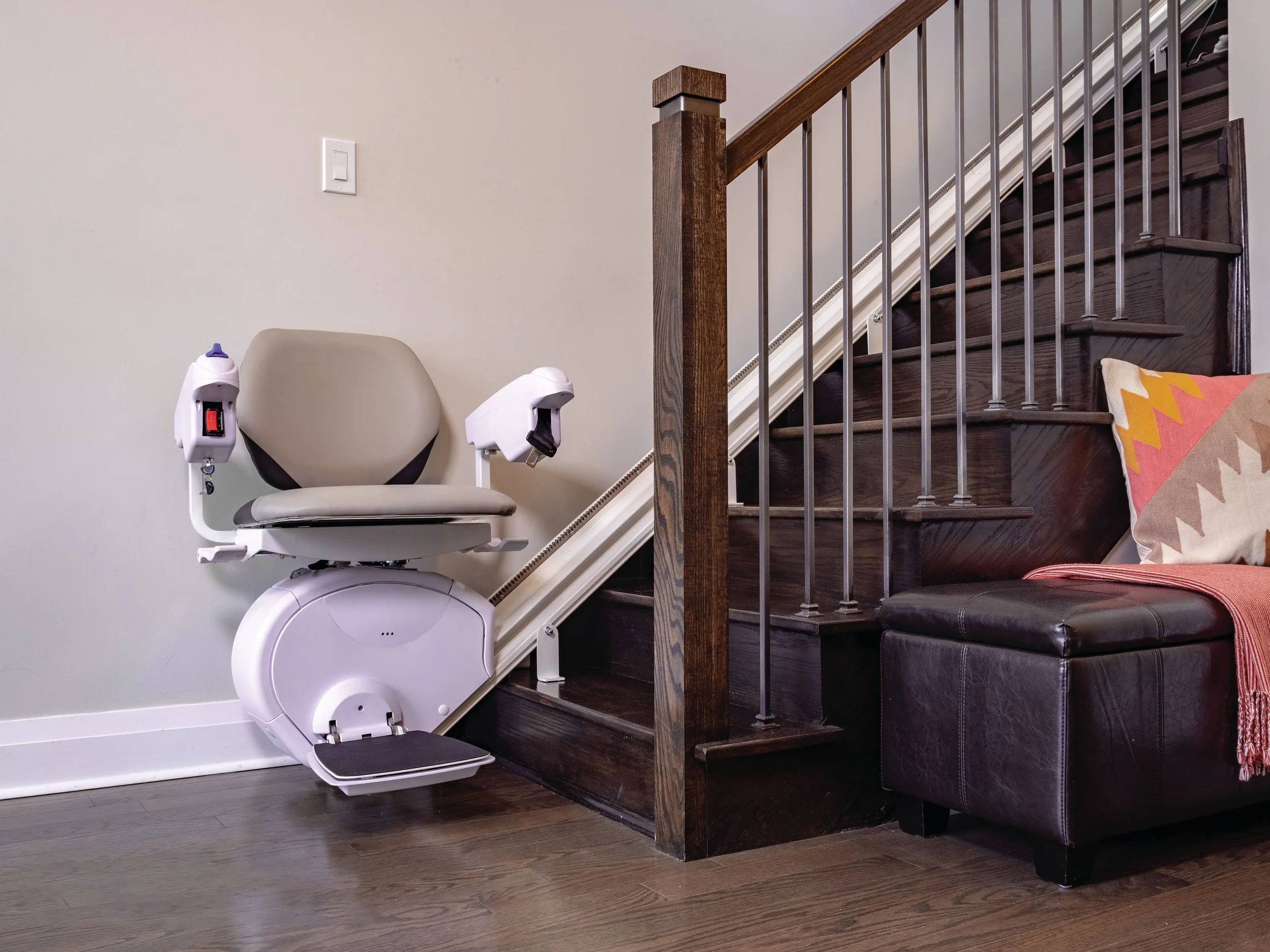 Silla de elevación para personas con discapacidad junto a la escalera de una casa, cerca de un sillón con cojines decorativos.
