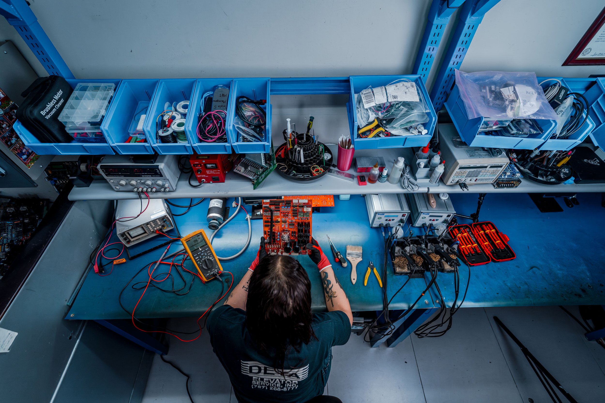 Persona trabajando en reparación de componentes electrónicos en un taller, con herramientas y dispositivos electrónicos organizados en mesas y estantes.