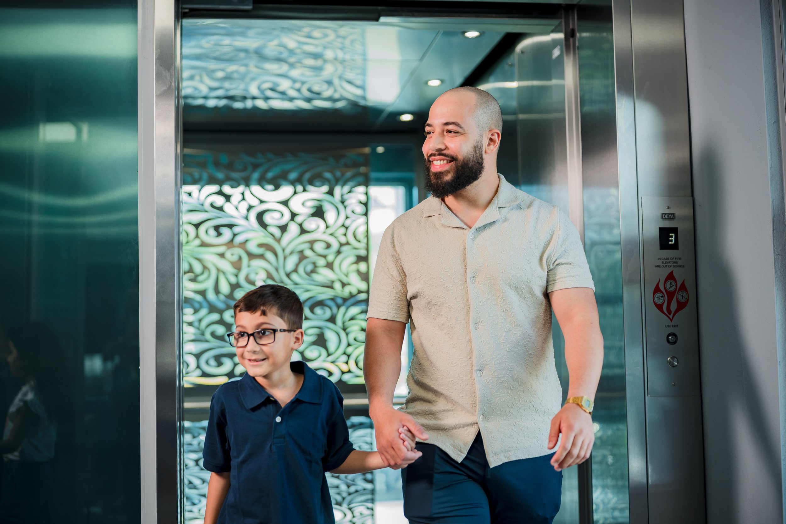 Hombre y niño con gafas sonriendo y caminando juntos en un ascensor, sosteniéndose de las manos.