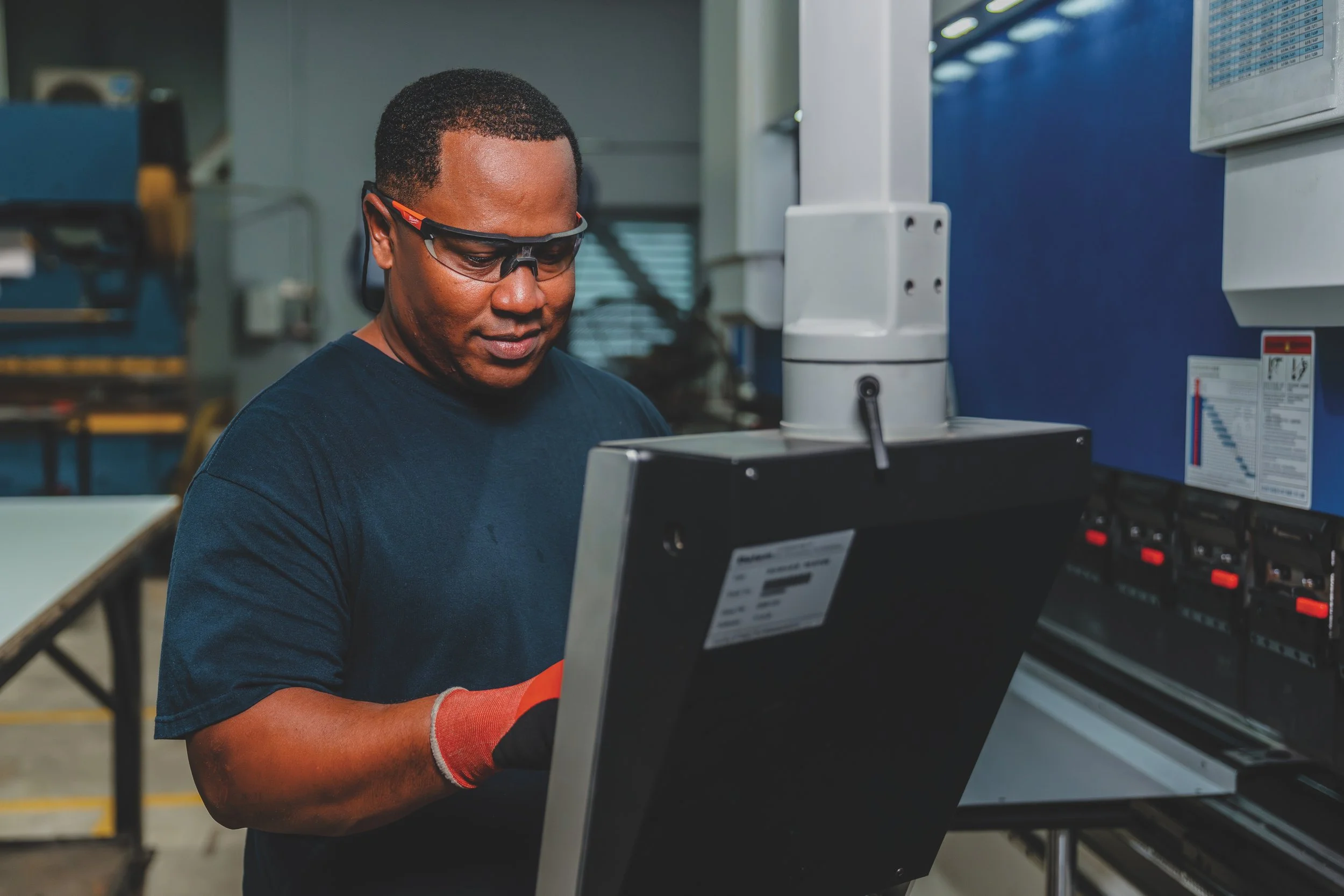 Hombre en un ambiente industrial o de manufactura, usando gafas de protección y guantes, trabajando con una máquina o equipo técnico.