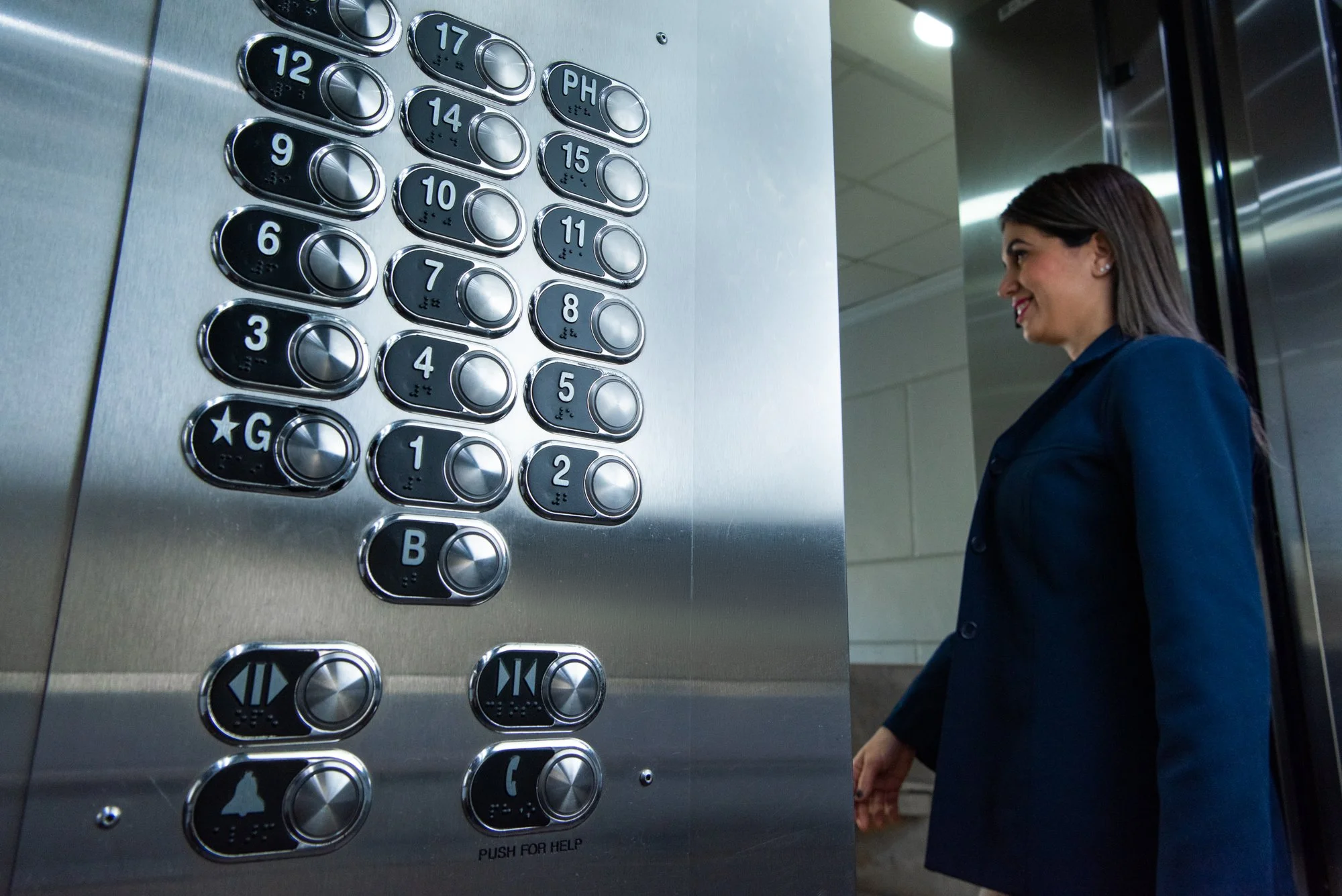 Una mujer sonriente en un traje azul de pie frente a un panel de botones de un elevador, listos para seleccionar un piso.