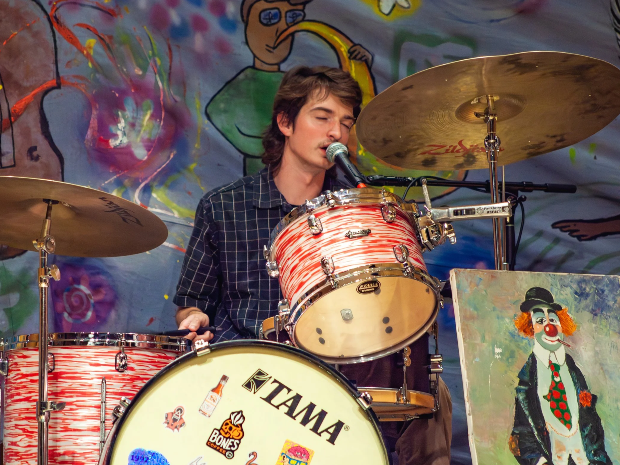 A young man with brown hair, wearing a dark plaid shirt, is singing into a microphone while playing drums. The drum set is red and white striped with stickers on the bass drum. Behind him is a graffiti-style backdrop with colorful abstract art, including a cartoonish character with glasses and a yellow beak, and a painted clown with orange hair and a red clown nose.