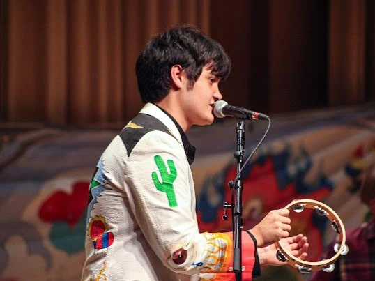 Young man singing into a microphone while holding a tambourine, on stage with a colorful backdrop.