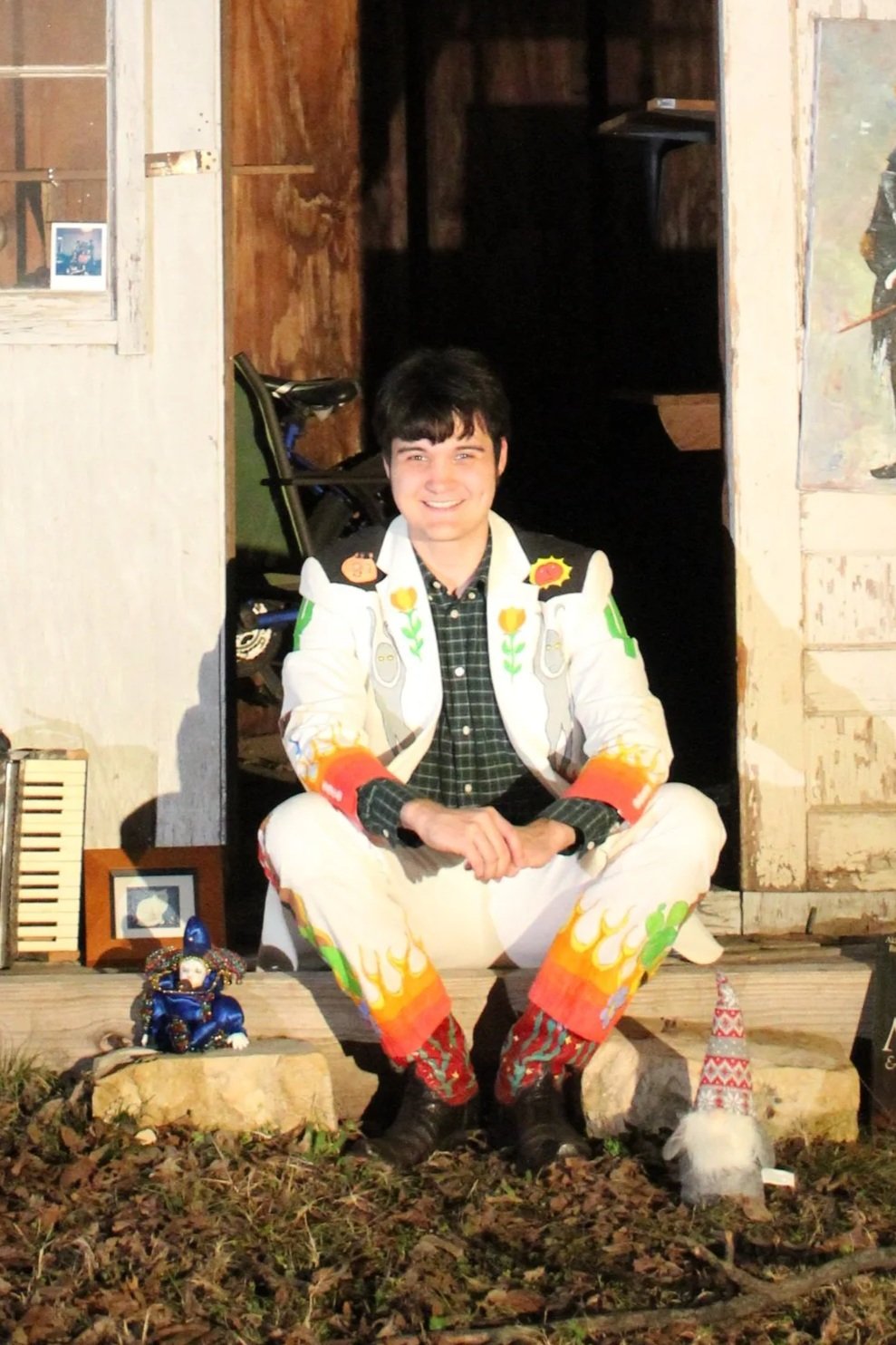 A young man with dark hair and a big smile, sitting cross-legged on the porch of a rustic wooden building. He's wearing a colorful, patterned jacket and pants, with festive designs. There are small decorative figures and objects around him, including a gnome and a doll, suggesting a cheerful or holiday setting.