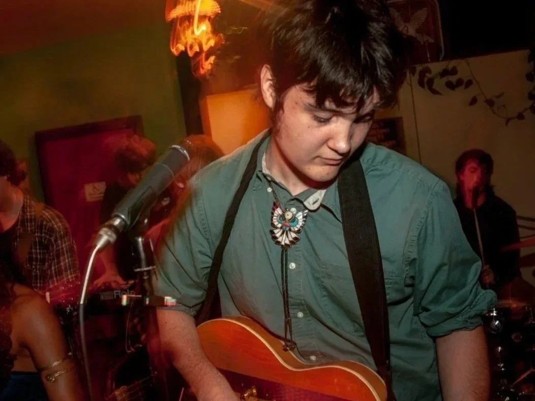 Young man with dark hair and wearing a necklace, playing an acoustic guitar in a dimly lit room with people around and a neon sign in the background.