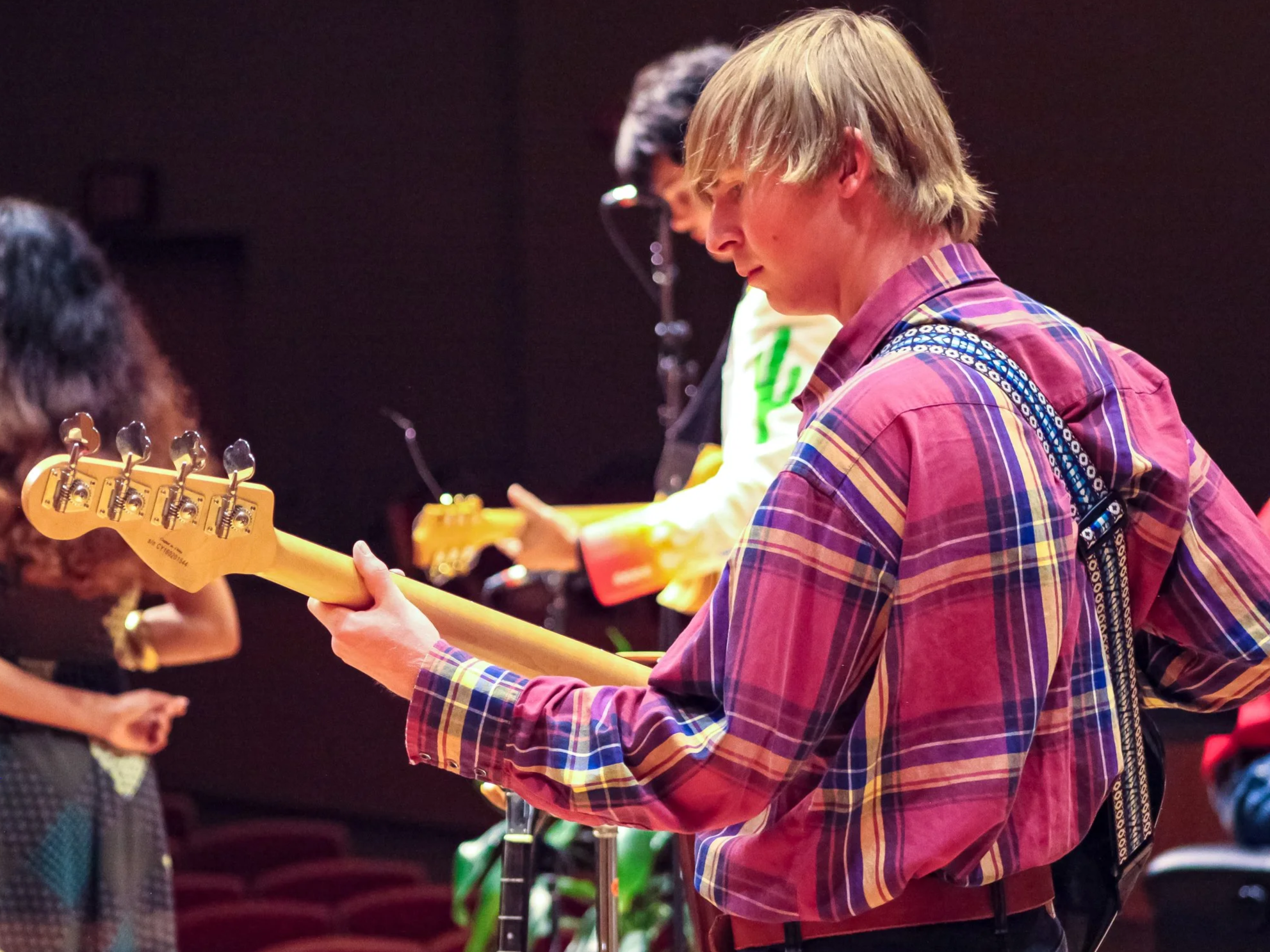 A person with blond hair wearing a colorful plaid shirt playing an electric guitar at an indoor music event.