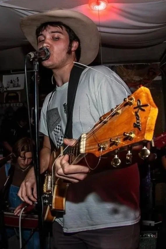 A man wearing a cowboy hat is singing into a microphone while playing an acoustic guitar with a unique headstock design in a dimly lit room, possibly a music venue or bar.
