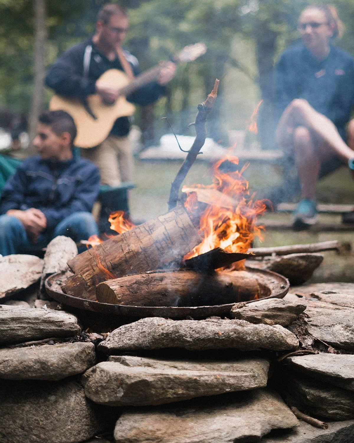 Burning campfire in focus, with group of people blurred in the background behind the fire