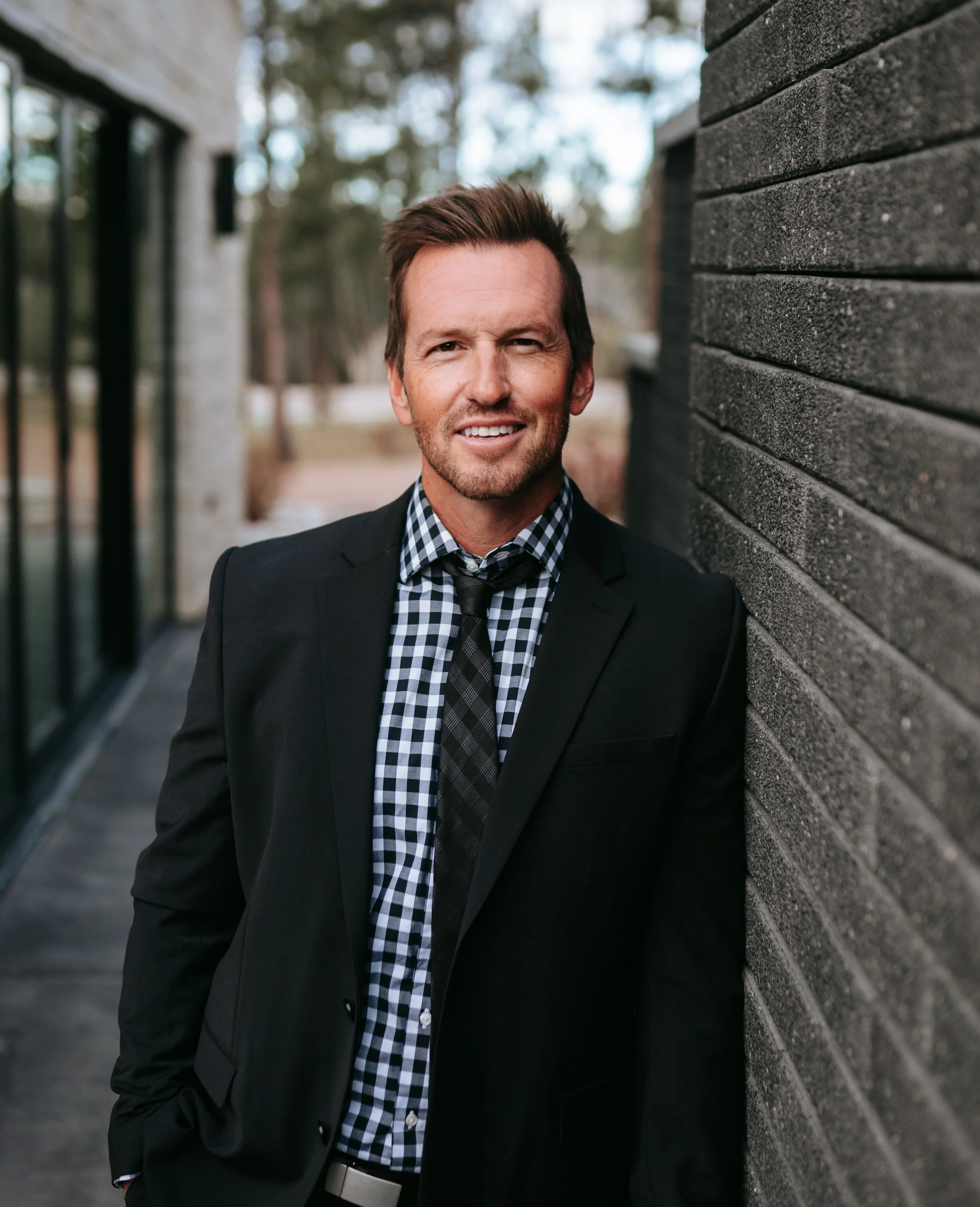 A man in a black suit with a checkered shirt and tie leaning against a dark brick wall outdoors, smiling at the camera.