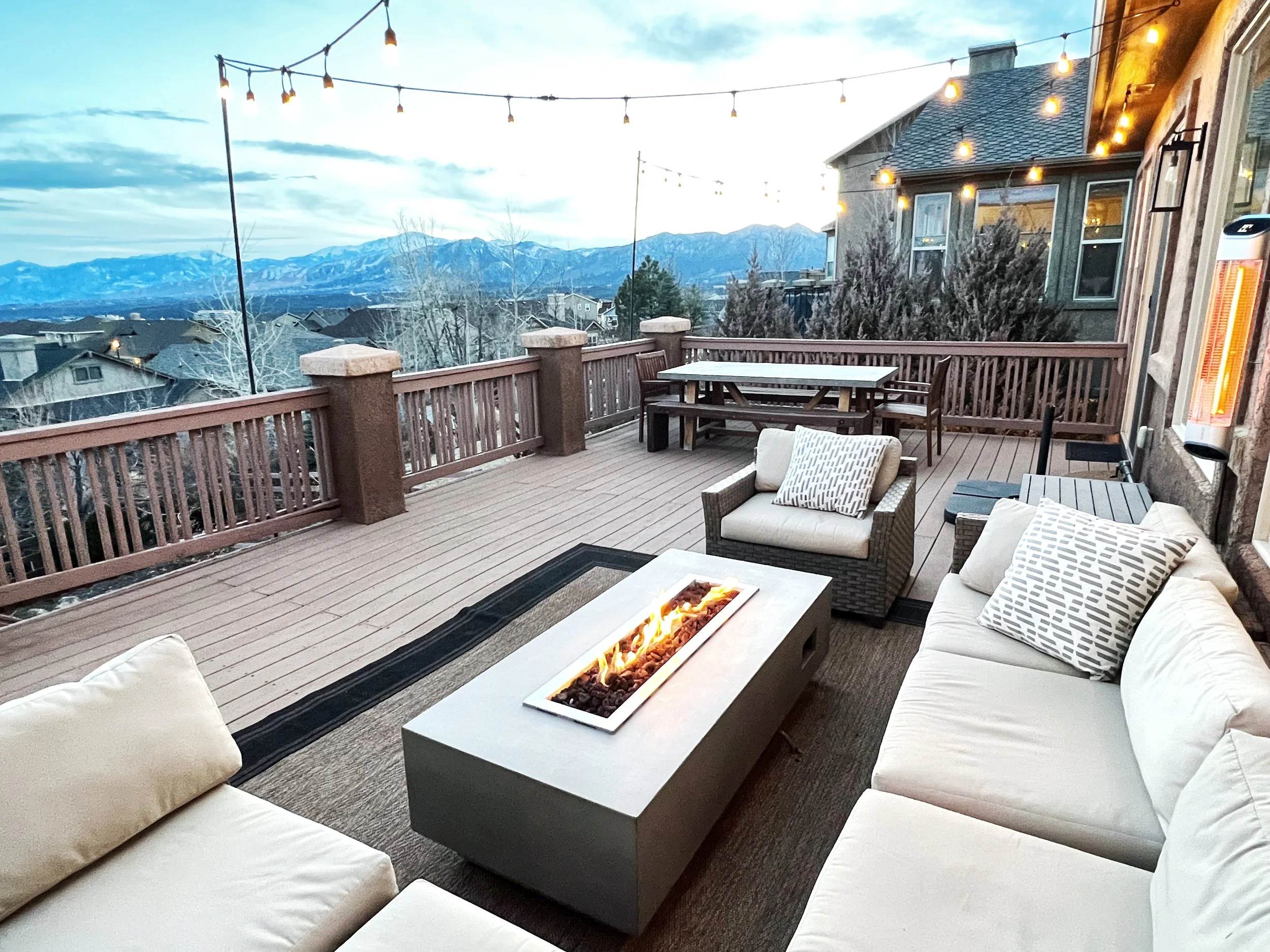 A spacious outdoor deck with a fire pit table, seating area with white couches and pillows, a dining table, and scenic mountain views in the background. String lights are hung overhead.