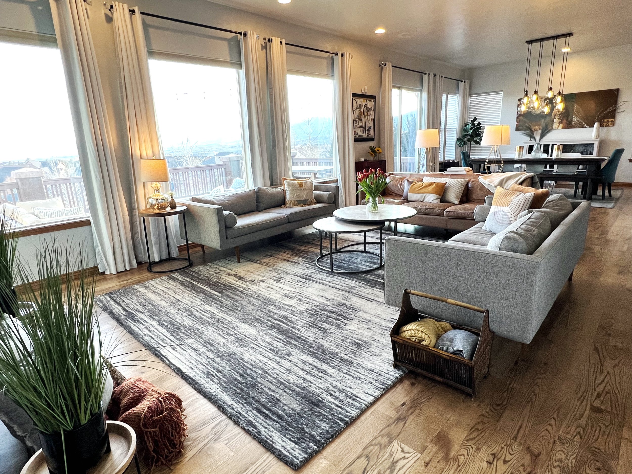 Bright living room with large windows, white curtains, and hardwood floors. Contains sofas, coffee tables, table lamps, and decorative elements including a vase of tulips.