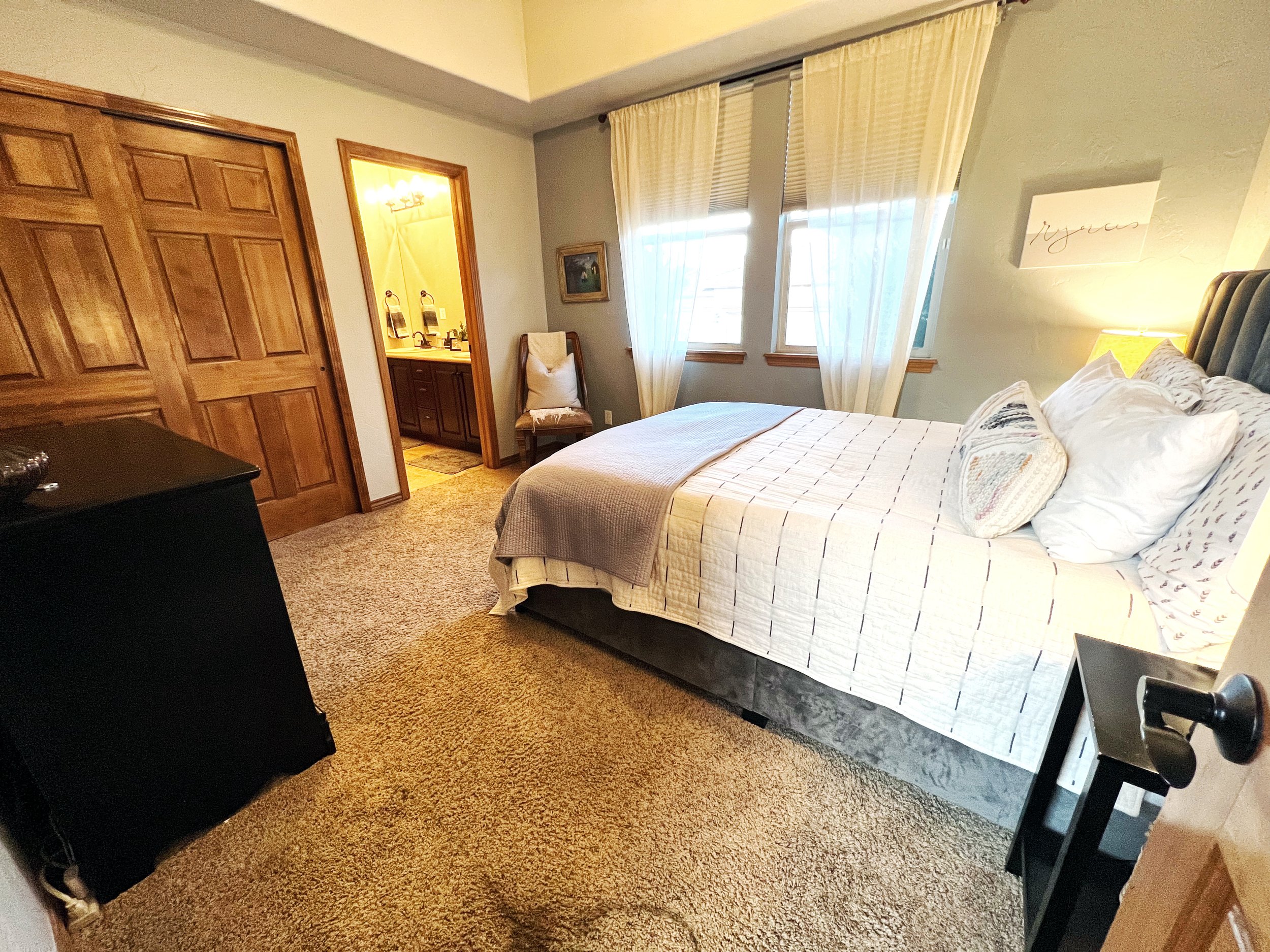 A cozy bedroom with a bed, window with curtains, a small bedside table with a lamp, and a bathroom visible through an open door.