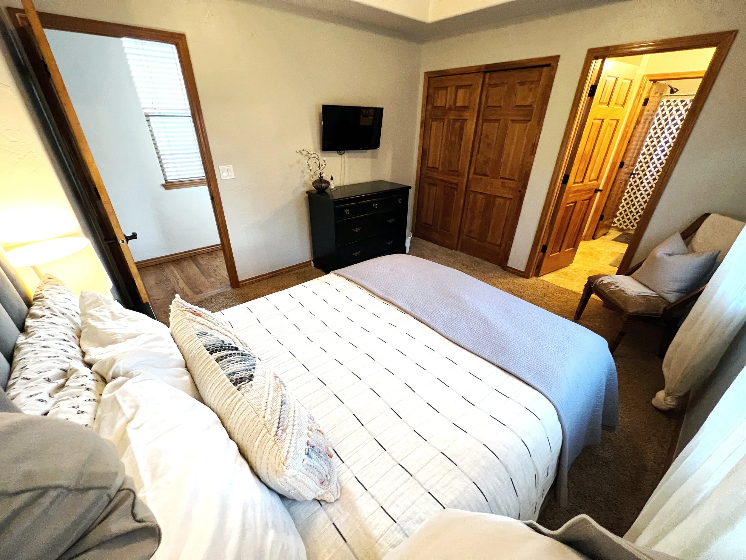 A bedroom with a bed in the center, covered with a white and black striped comforter and pillows. There is a small black dresser with a decorative vase and branches, a mounted flat-screen TV, a wooden closet, and an open doorway leading to a bathroom