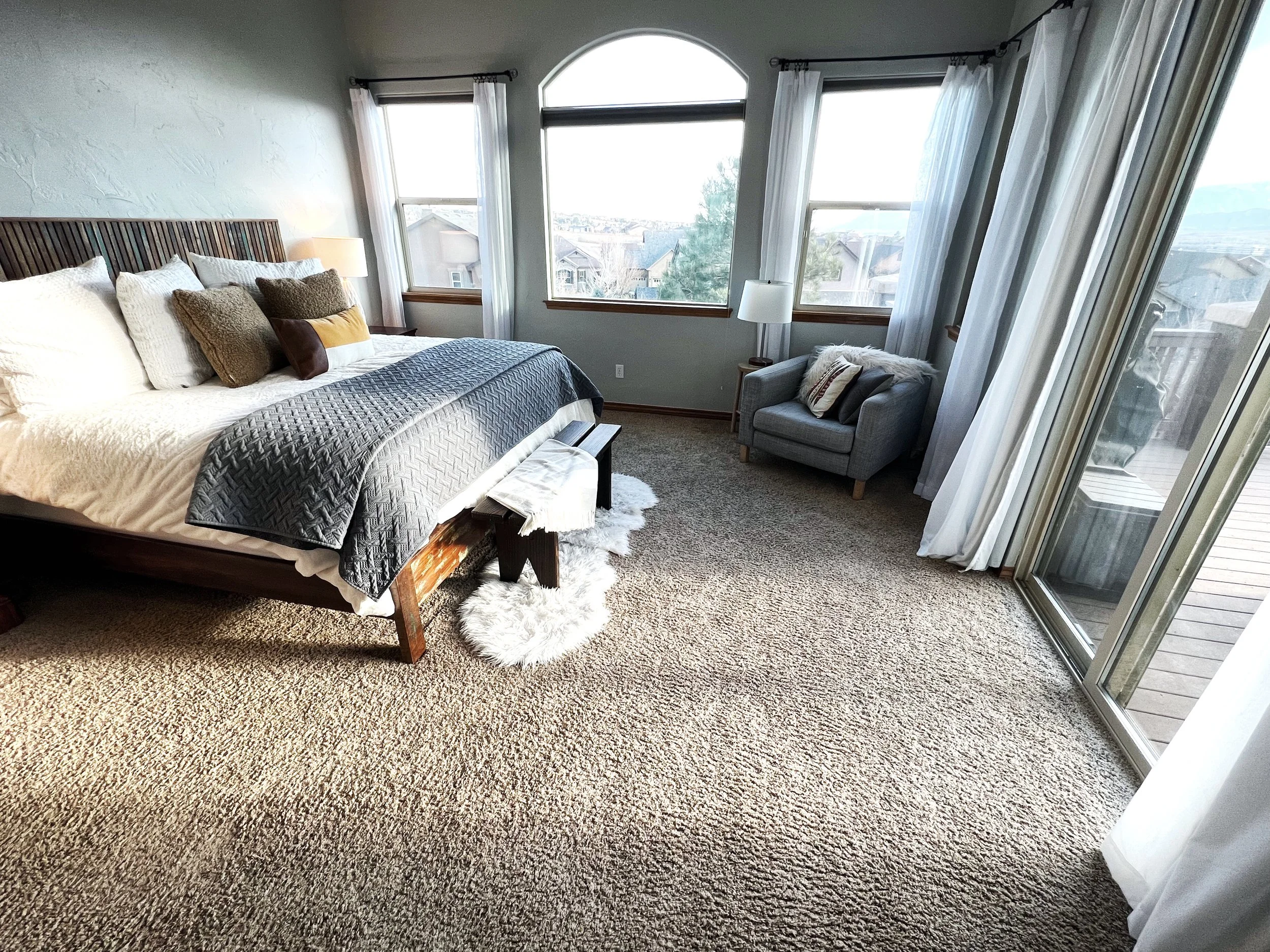 A cozy bedroom with a large bed, pillows, a bench at the foot of the bed, a small gray armchair, a white furry rug, and large windows with white curtains letting in natural light.