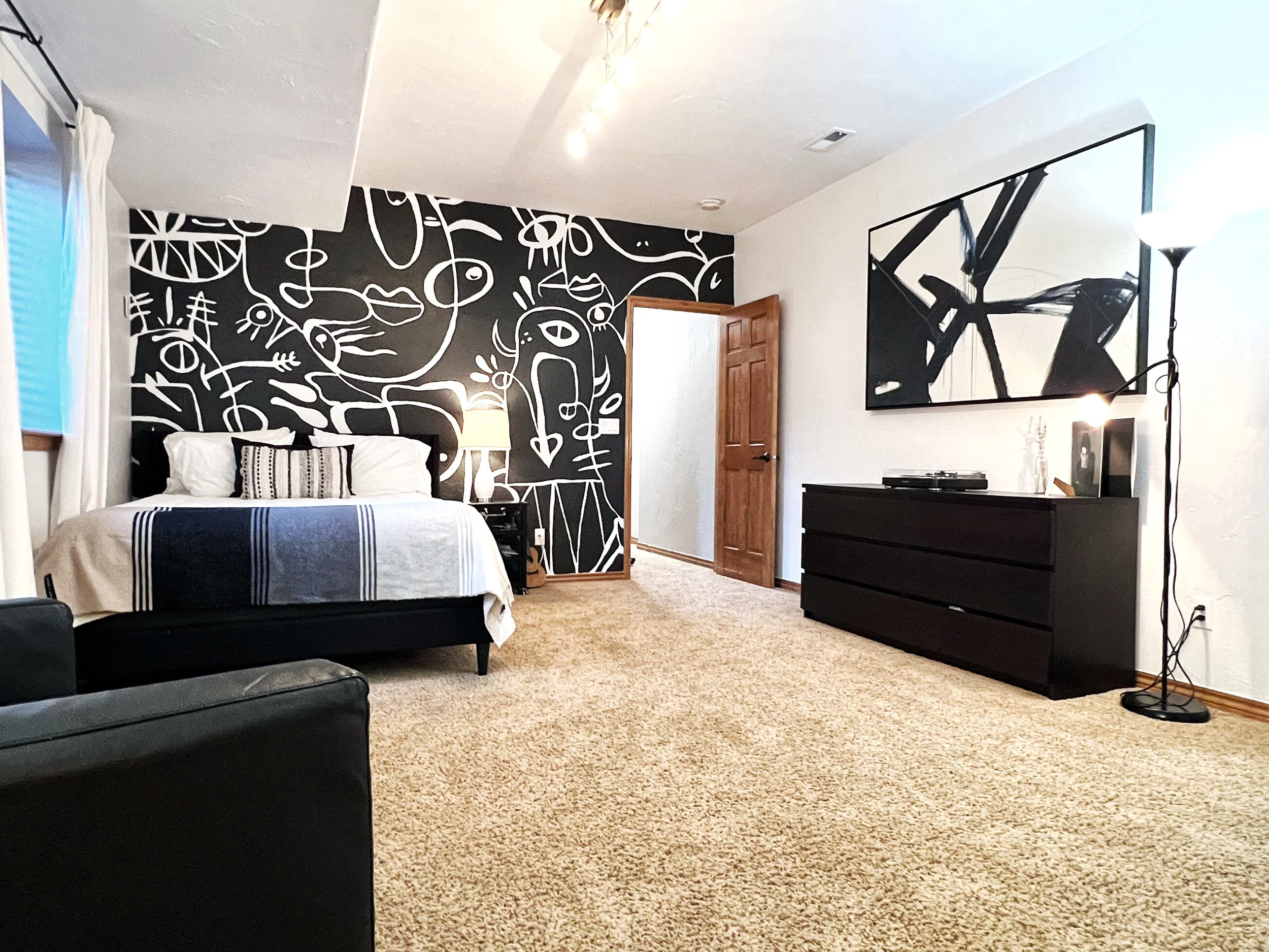 A modern bedroom with a black and white abstract art wall behind the bed, a black dresser with a large mirror above, a floor lamp, a bed with striped bedding, and a beige carpeted floor.