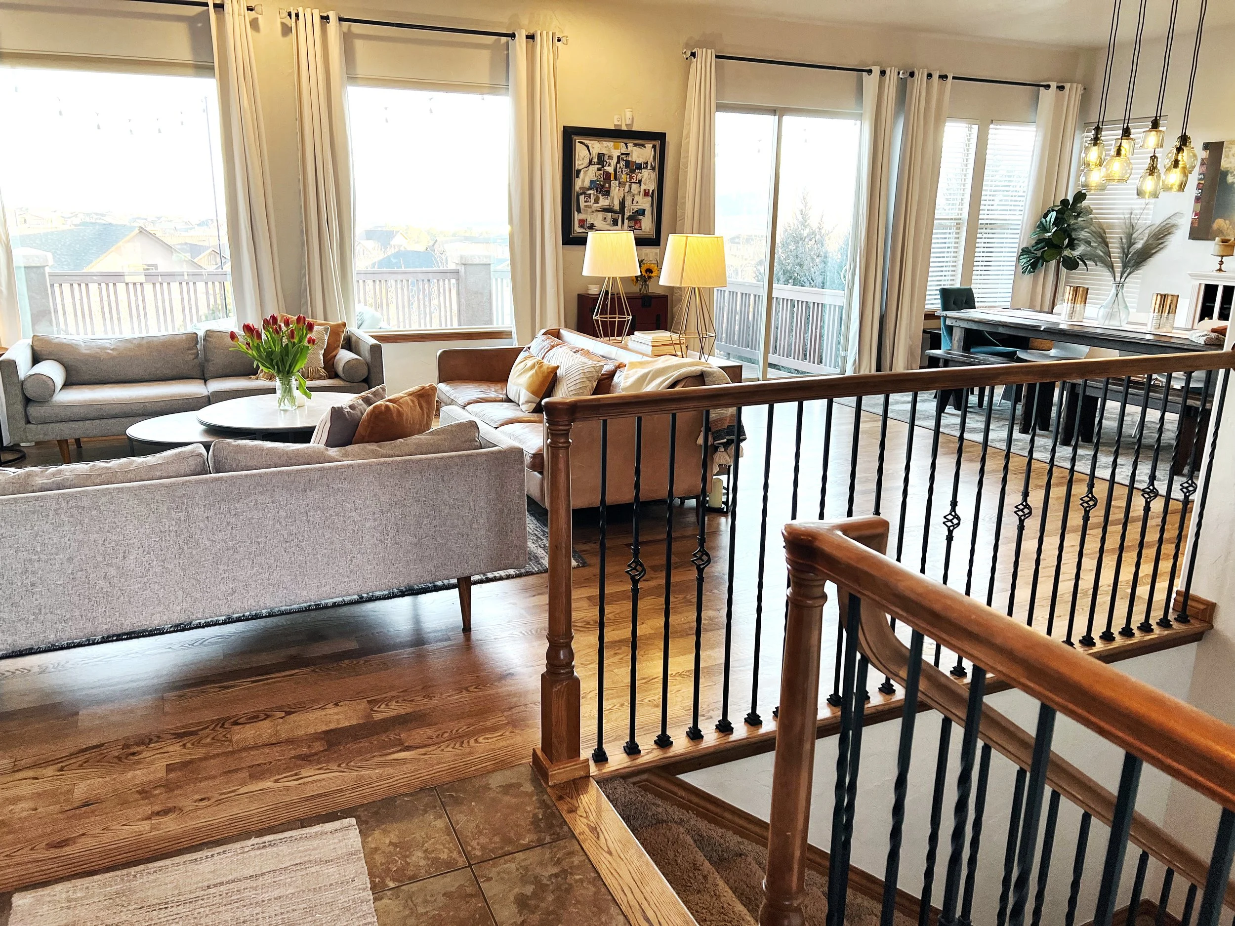Bright living room featuring multiple couches, a round coffee table with a vase of red tulips, large windows with cream curtains, a wooden staircase railing, and a dining area with a table, chairs, and decorative plants.