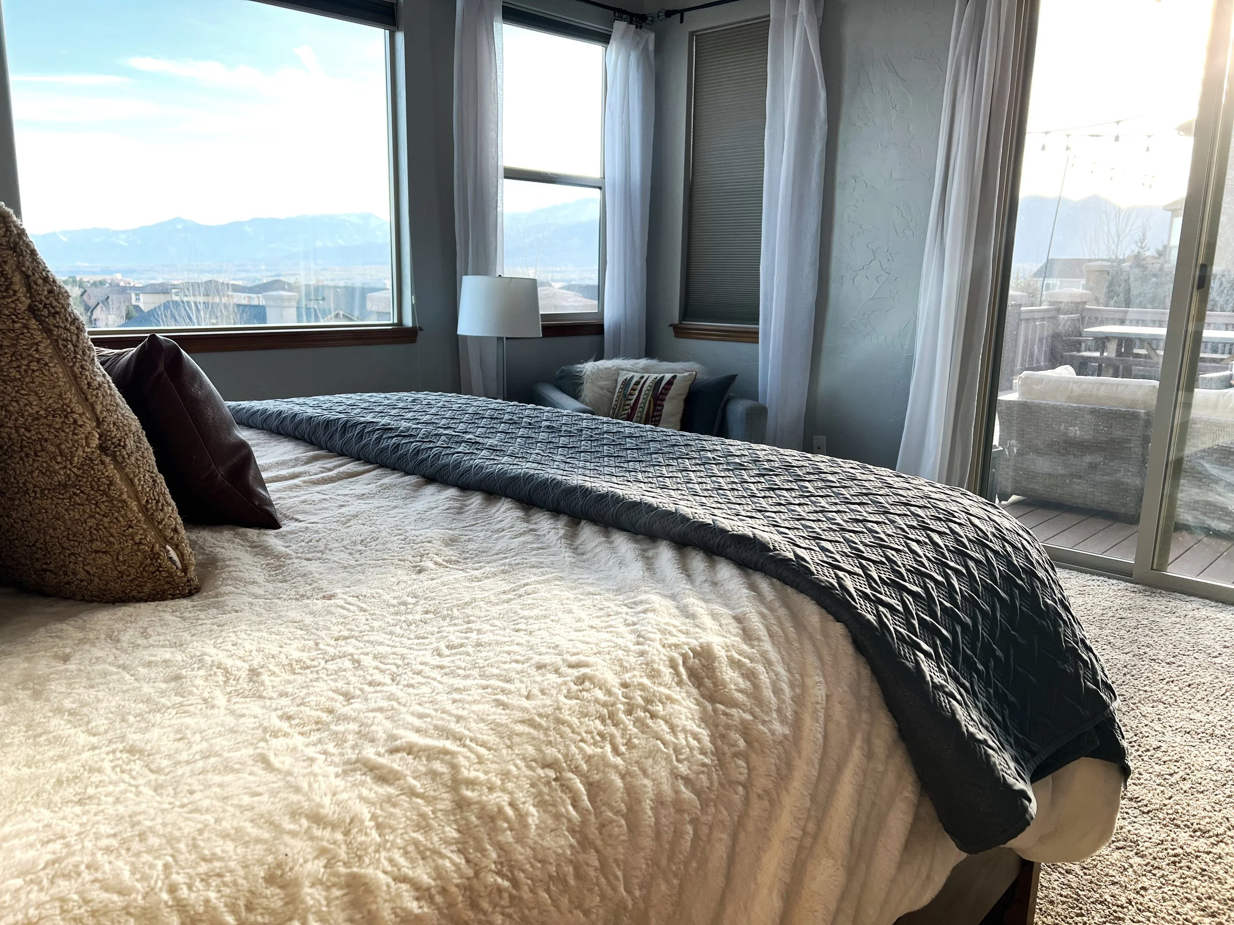 A cozy bedroom with a large bed covered in a textured cream blanket and dark pillows, overlooking a mountain view through two windows and a sliding glass door leading to a balcony with outdoor furniture. Light curtains frame the windows and door.
