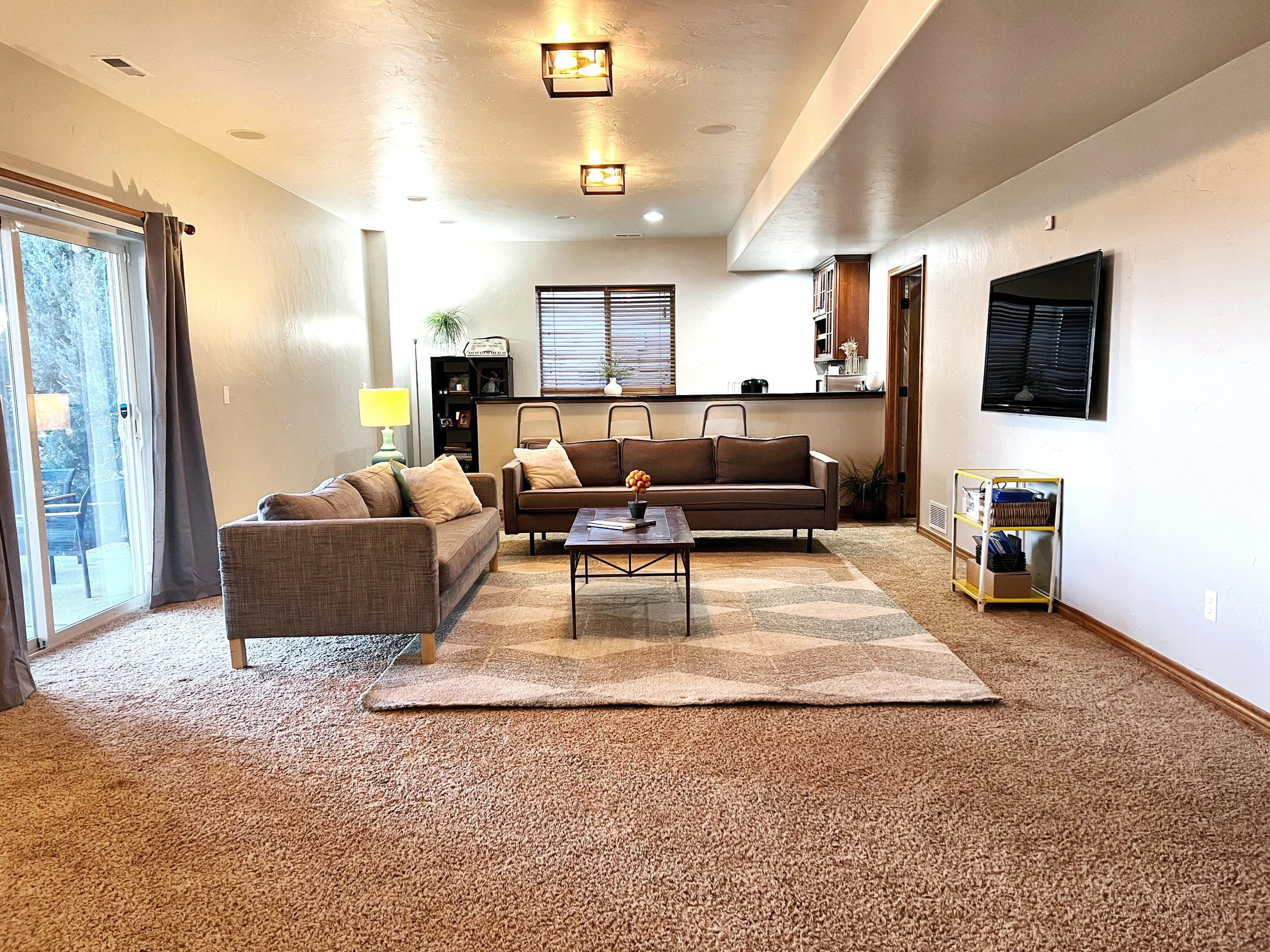 Living room with two sofas, a coffee table, a television on the wall, a sliding glass door, a small white bookshelf, and a window with blinds. The room has beige walls and carpeting, with a rug in the center.