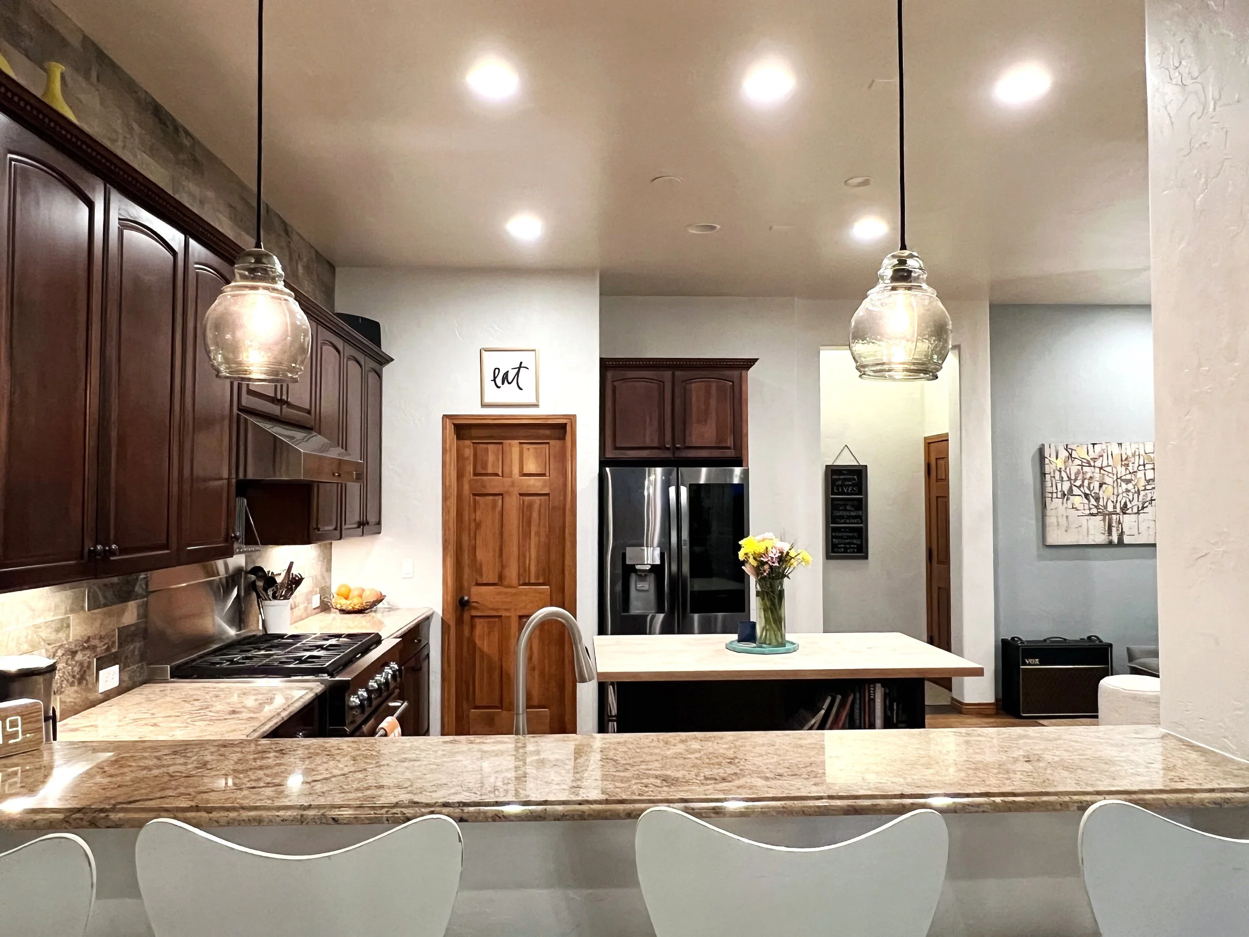 View of a modern kitchen with dark wood cabinets, a marble countertop, and a kitchen island with a vase of flowers. Recessed lighting and pendant lights hang from the ceiling. There are stainless steel appliances, a wall art sign that says 'eat', and