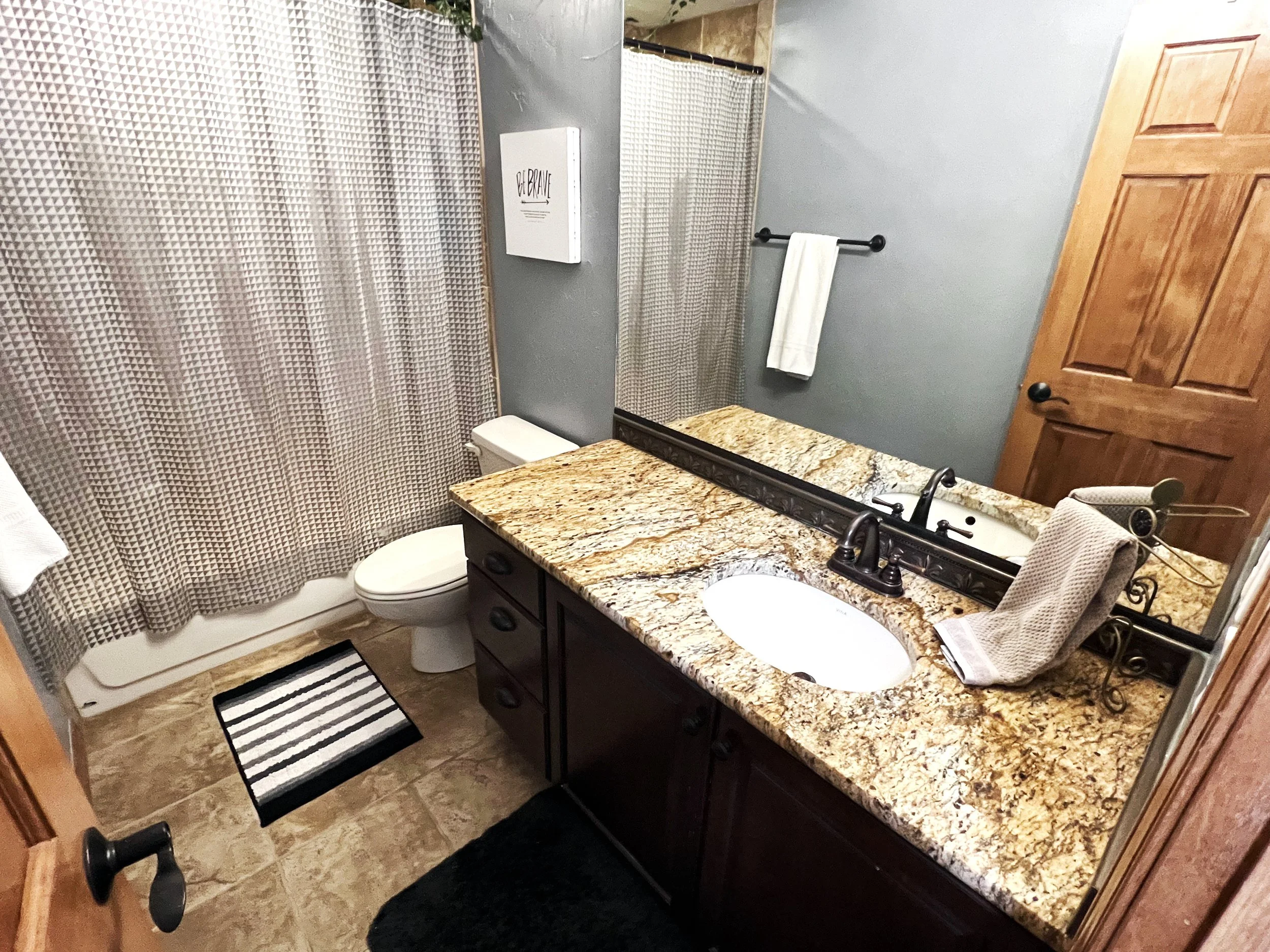 The image shows a bathroom with a granite countertop vanity, a mirror, a toilet, a shower with a patterned curtain, a towel on a black pole, a wooden door, and a striped bath mat on the floor.