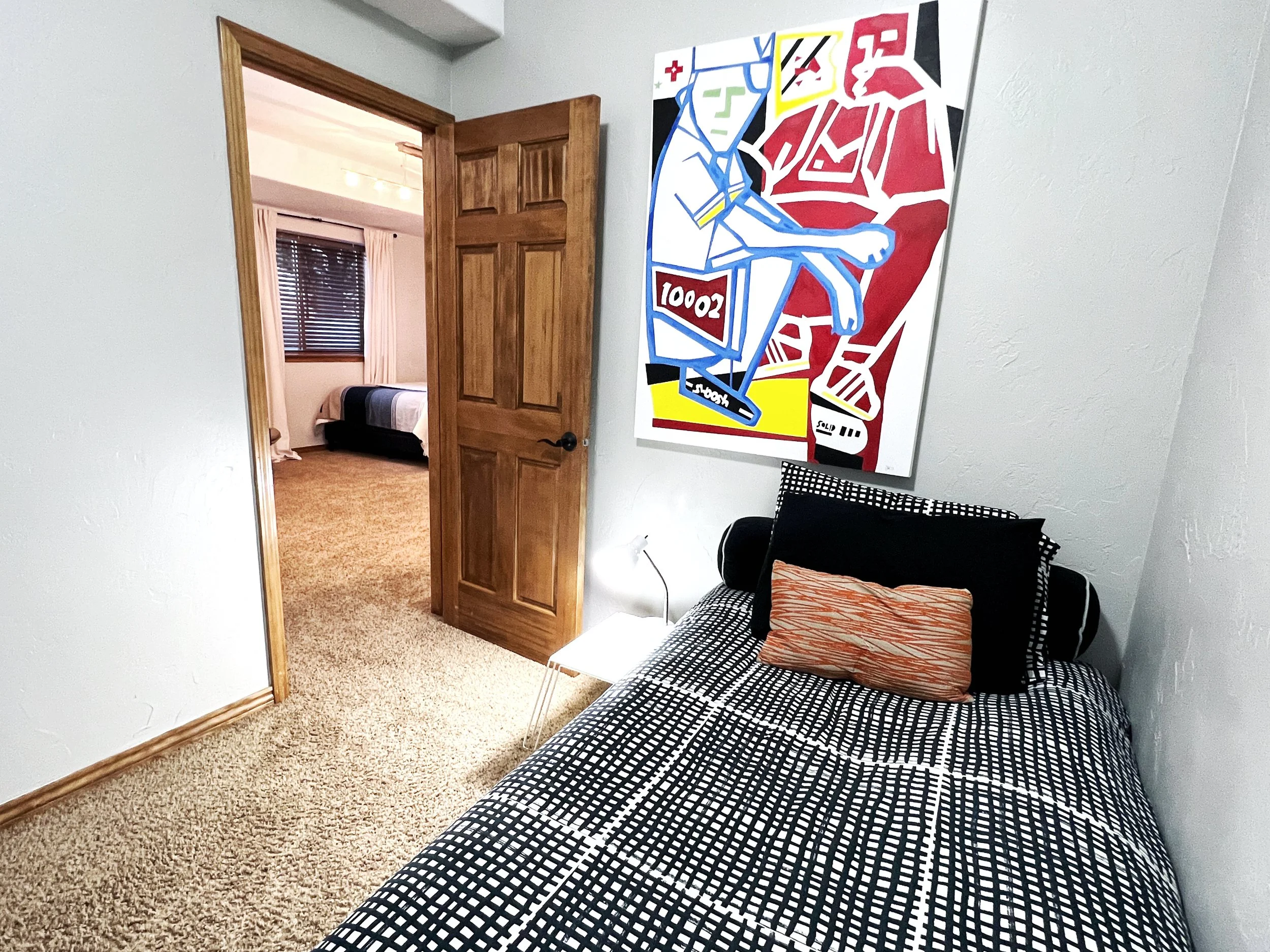 A bedroom with a single bed, black and white patterned bedding, a striped pillow, and a large colorful abstract artwork on the wall. The door is open, revealing another bedroom with a window and beige carpet.