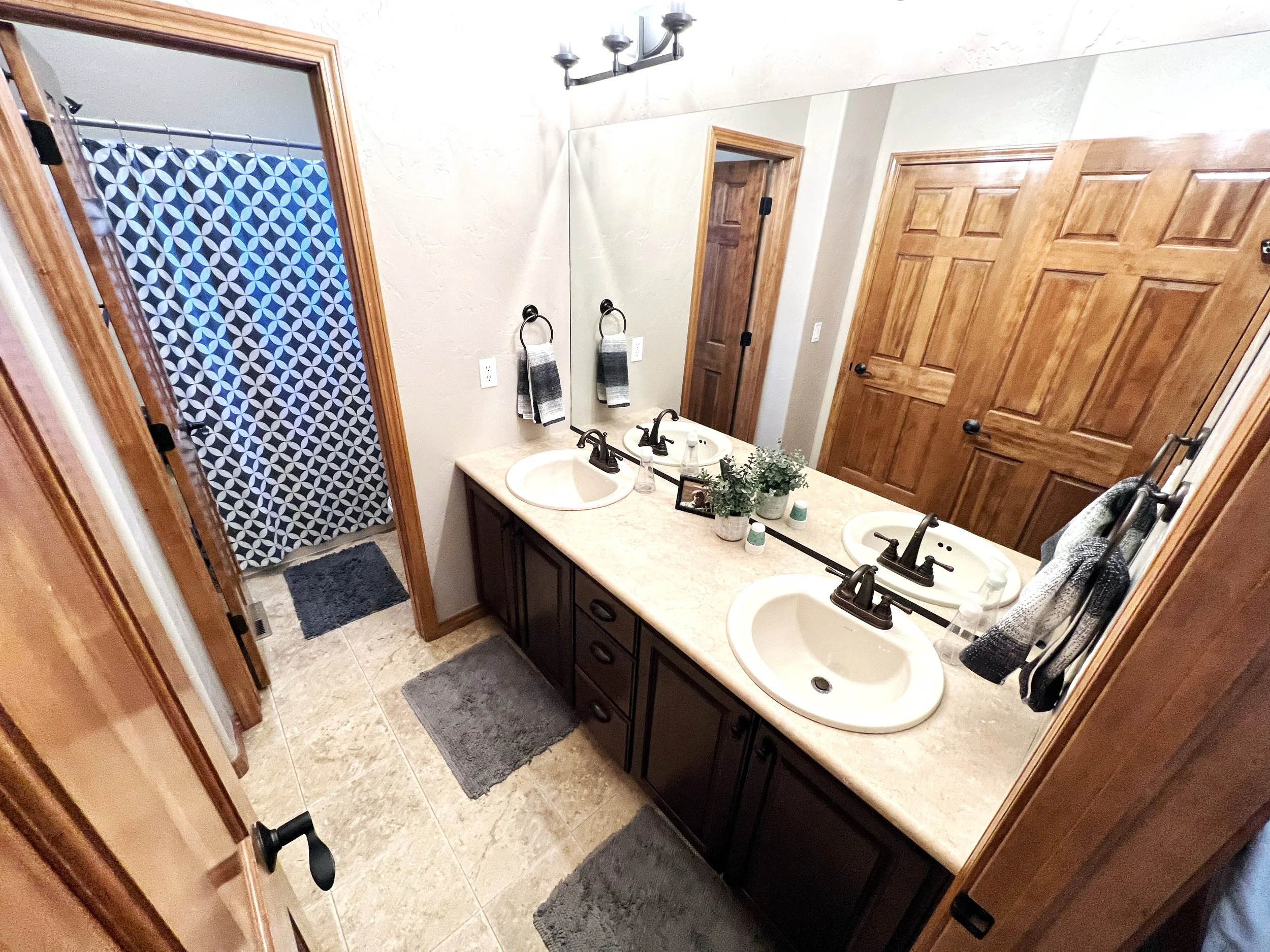 Bathroom with double sink vanity, large mirror, wooden cabinets, towel racks, decorative items, and a shower with geometric-patterned curtain.