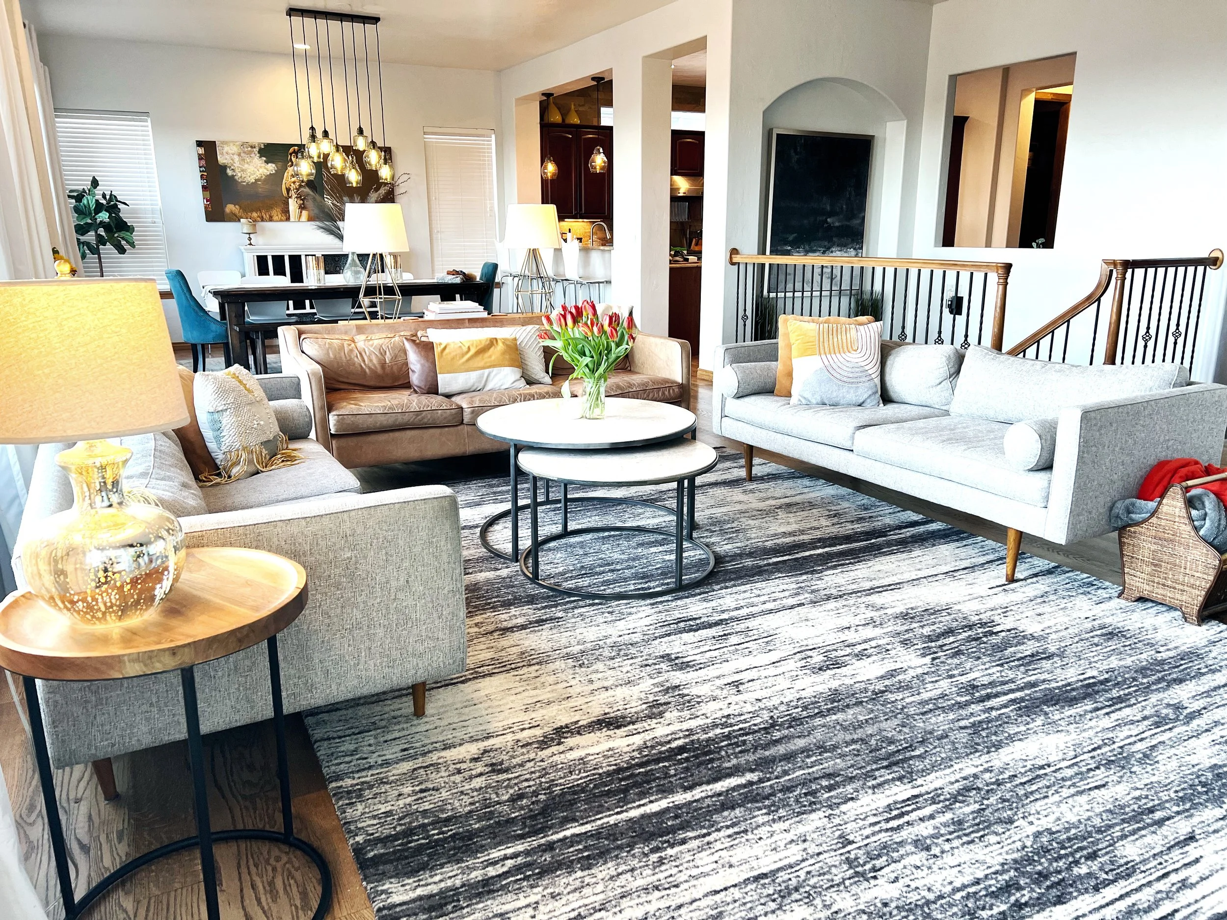 Open-concept living room with three sofas, two white and one brown, arranged around two nested round coffee tables with a vase of red tulips. A side table with a golden lamp is in the foreground. The background features a dining area with a black tab
