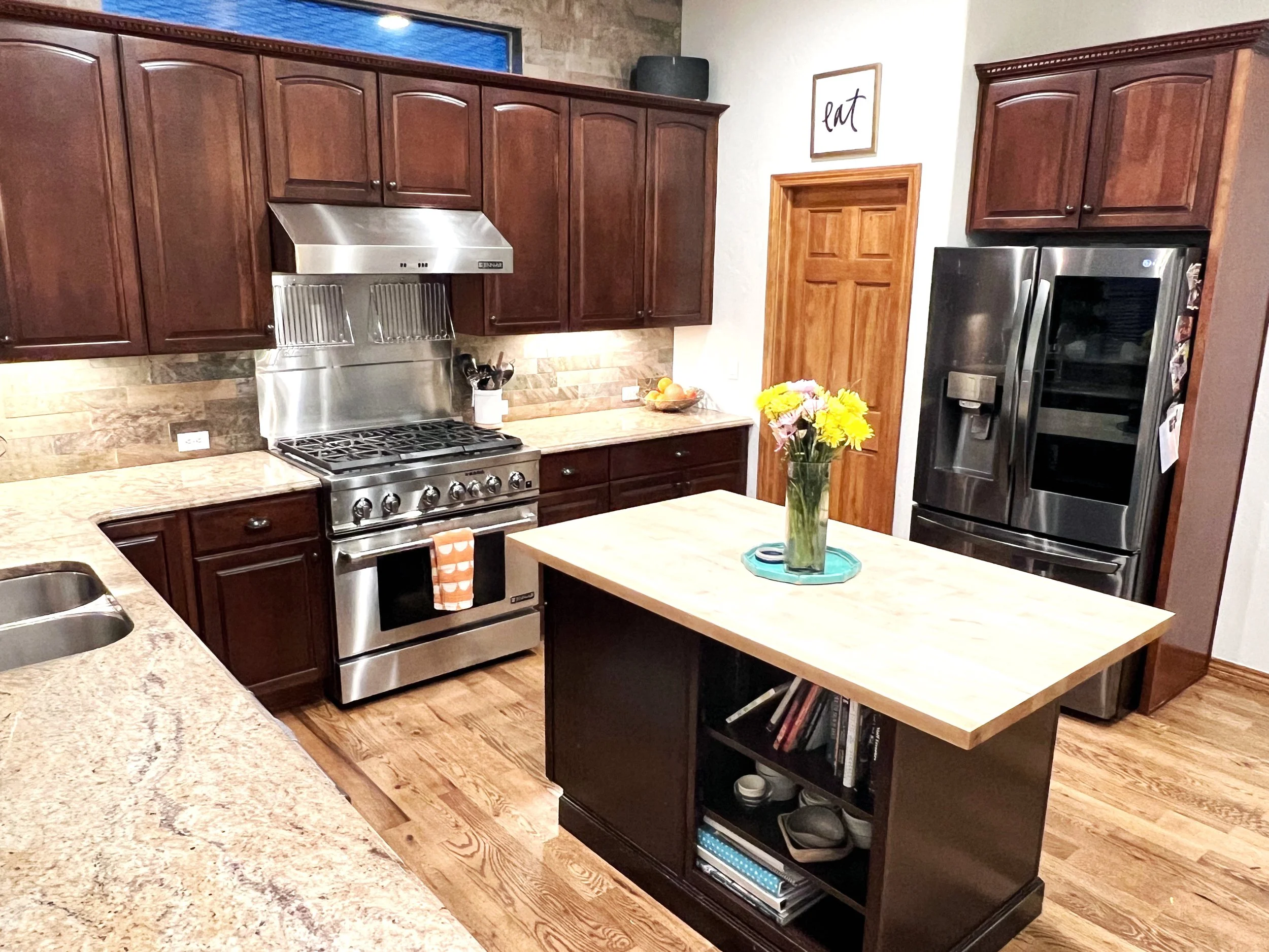 Modern kitchen with dark wood cabinets, stainless steel appliances including a refrigerator and a stove, a kitchen island with books and decor on the shelves, a vase of yellow flowers on the island, and a sign that says 'Eat' on the wall.