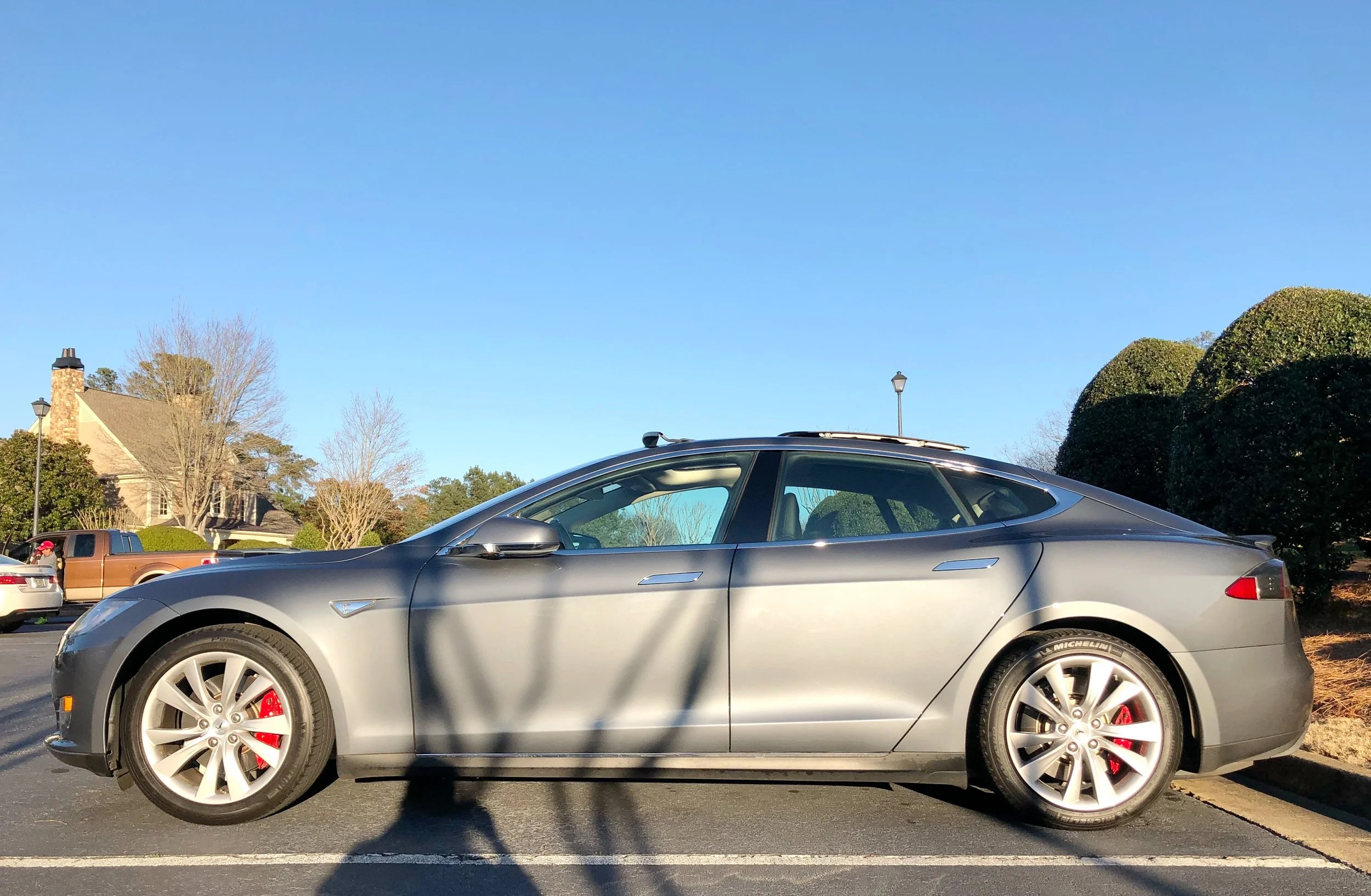 A silver Tesla Model S parked in a parking lot with a suburban neighborhood background, clear blue sky, and trees.