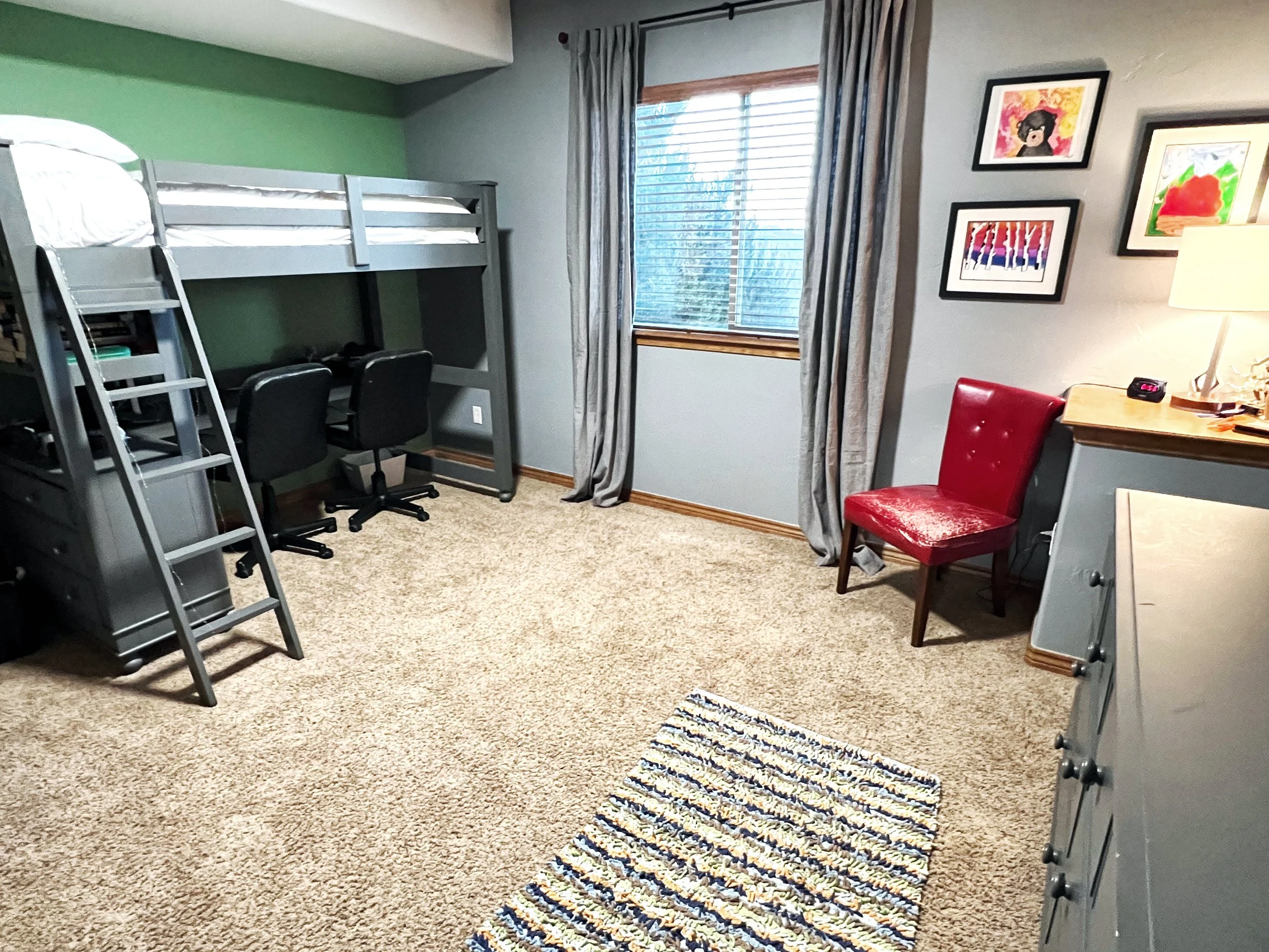 A bedroom with a loft bed, two chairs, a dresser, a red upholstered chair, artwork on the wall, a window with curtains, a lamp, and a striped rug on beige carpet.