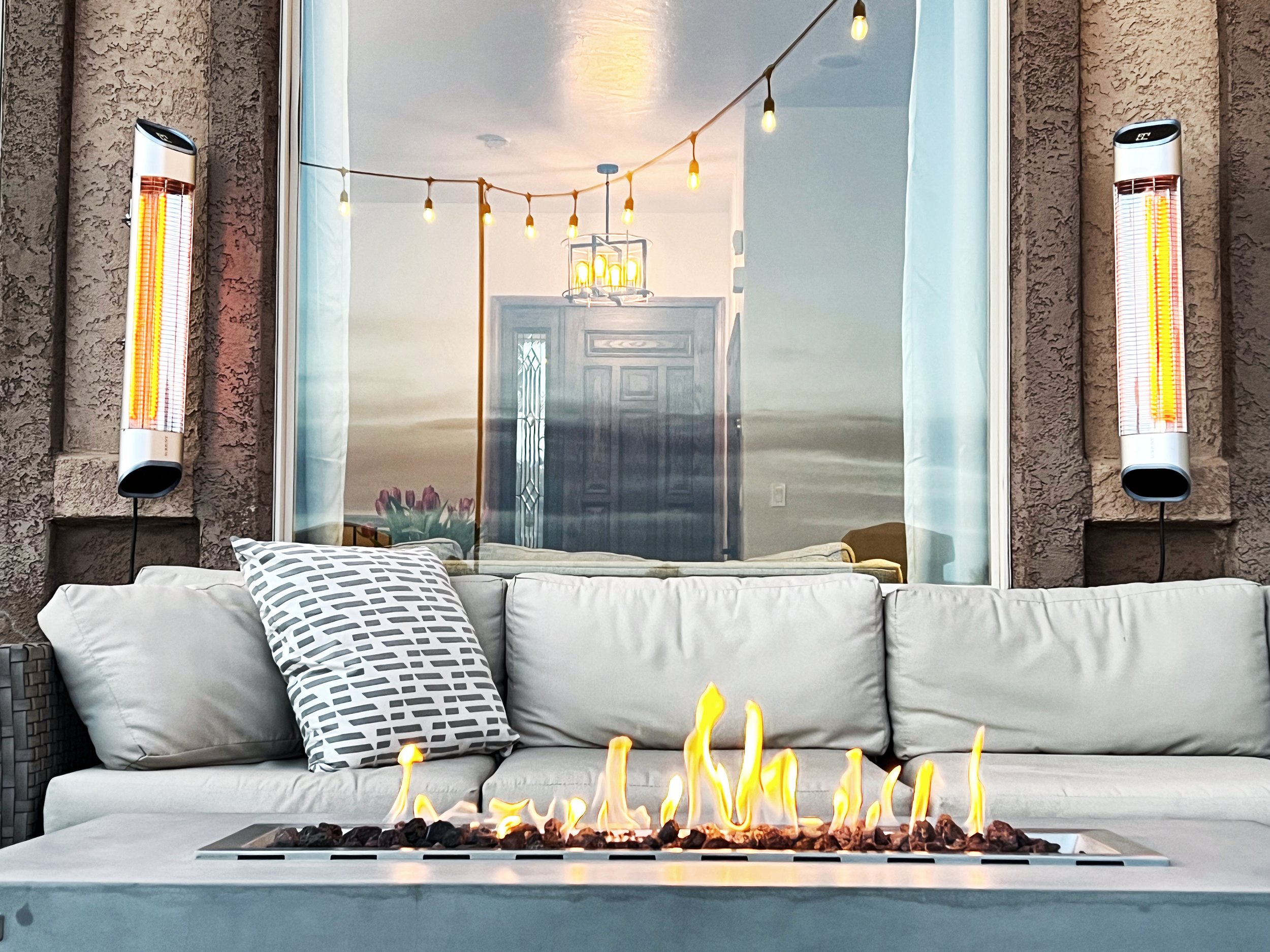 Outdoor patio with a gray cushioned sofa, decorative pillows, and a linear gas fire pit with flames. Behind the sofa, there are two wall-mounted heaters, a reflective glass window, and string lights hanging above. Reflections of a front door and pati