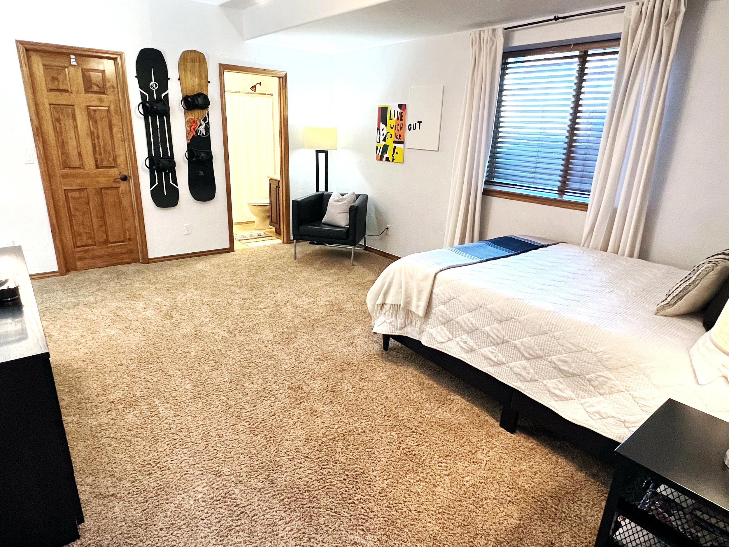 A cozy bedroom with a single bed near a window with closed blinds and cream curtains, a black armchair with a pillow, promotional posters on the wall, and a door leading to a bathroom with a toilet. There are two snowboards mounted on the wall.