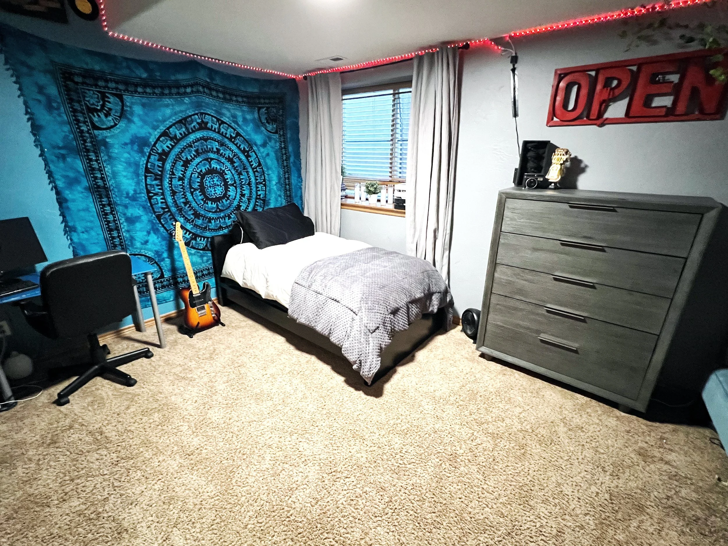 A bedroom with a bed, guitar, desk, and dresser. The wall has a large blue tapestry and a red "OPEN" sign. There's a window with white curtains and blue blinds, decorated with small plants and a radio. The floor is carpeted in beige.