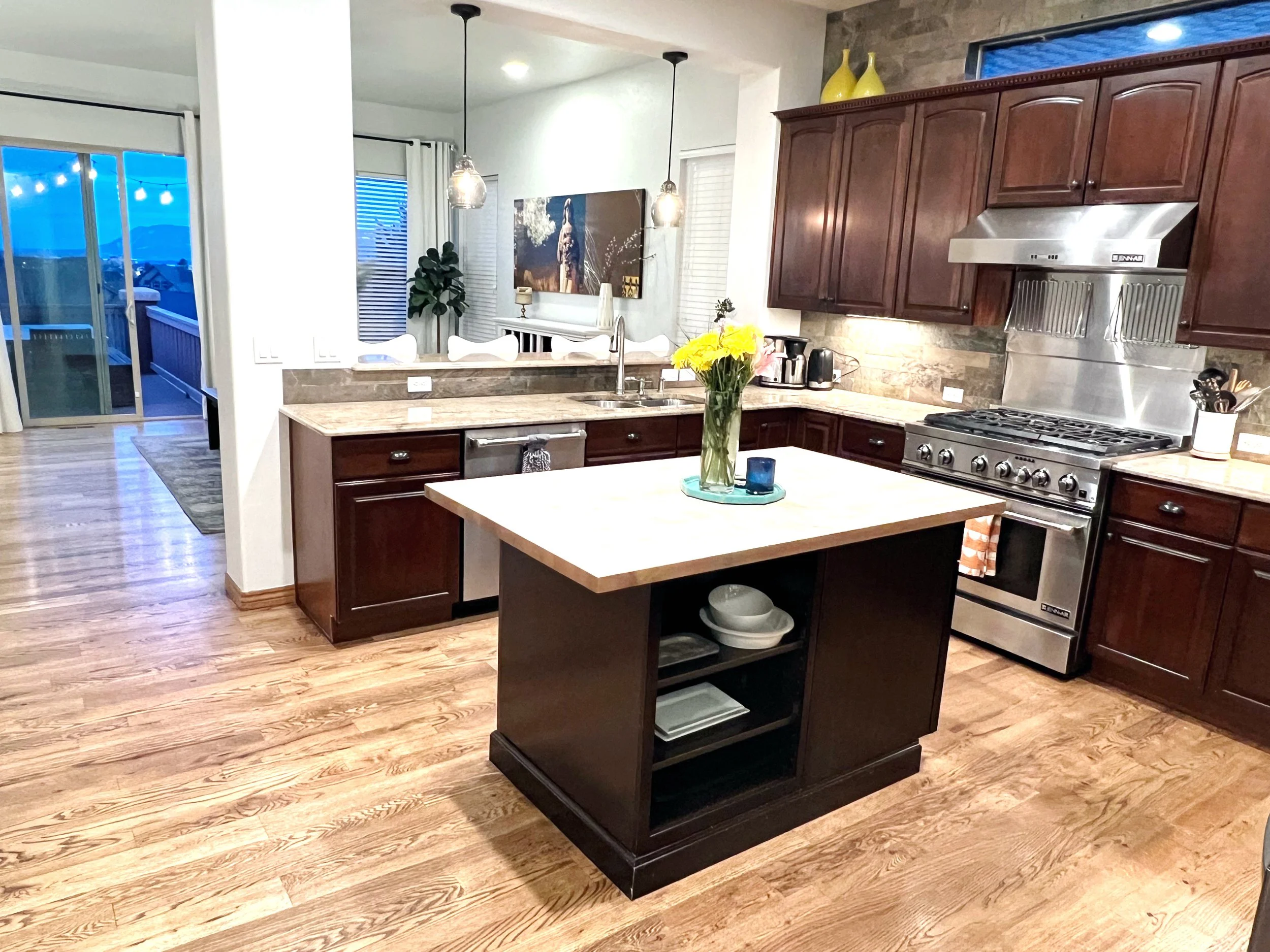 Modern kitchen with dark wood cabinets, stainless steel stove, and a small island with open shelves, decorated with a vase of yellow flowers, in a home with hardwood floors and a view into the living area.