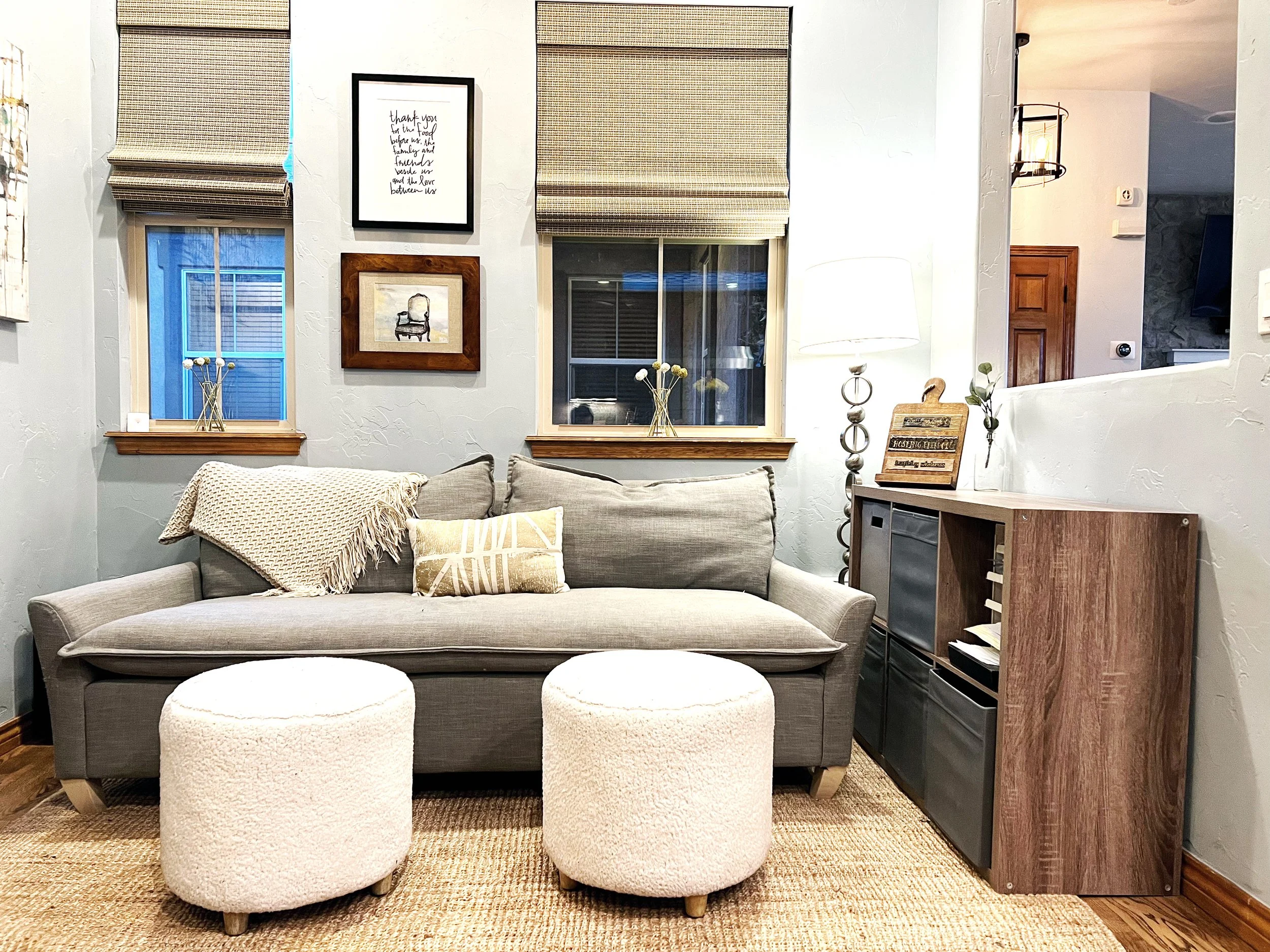 Living room with gray sofa, two white ottomans, wall art, windows with blinds, lamp, and wooden console