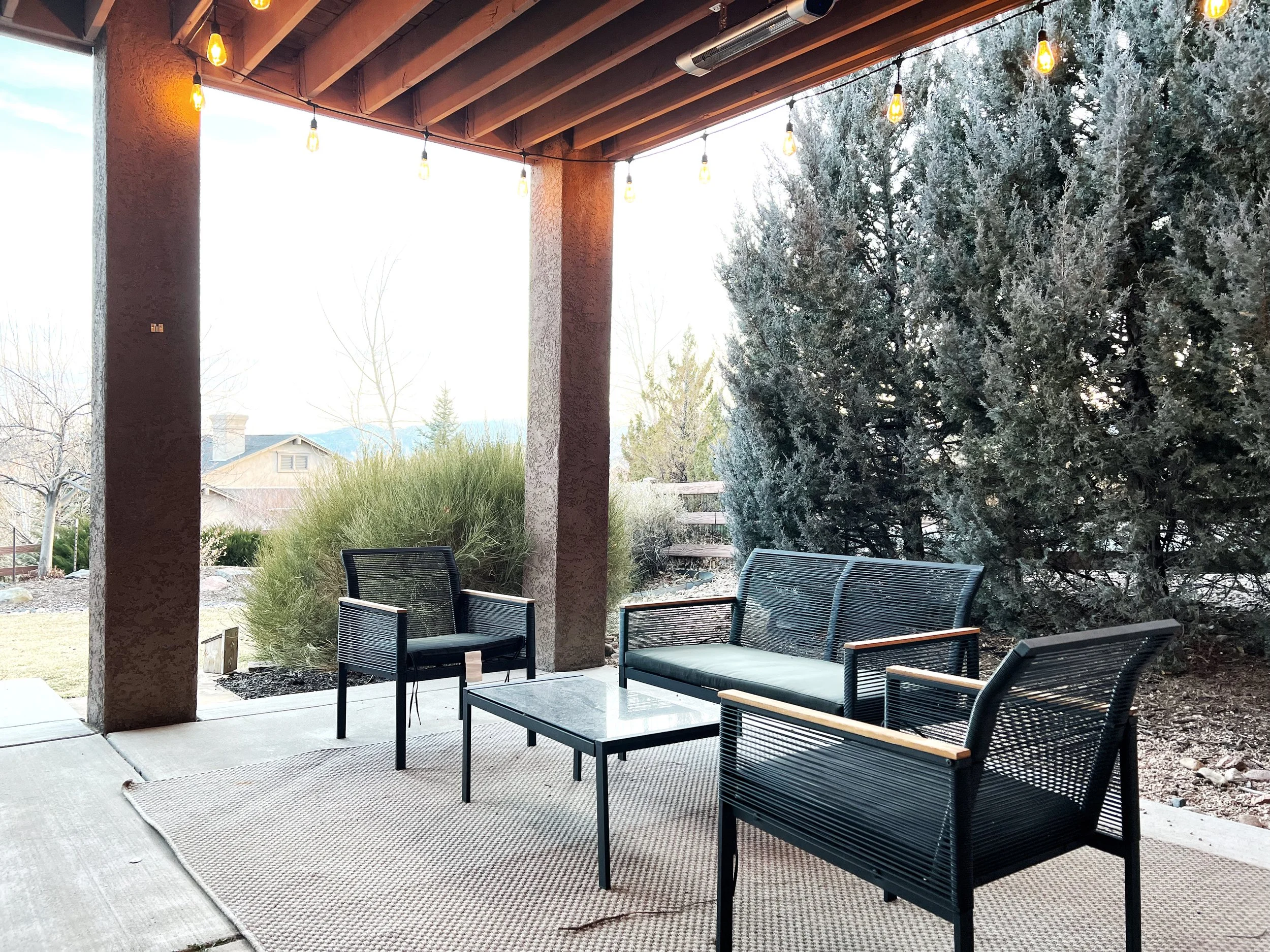 Outdoor patio with black outdoor furniture, including two armchairs, a loveseat, and a glass coffee table, on a beige rug. String lights hang from a wooden ceiling, and a large evergreen bush and other plants are in the background.