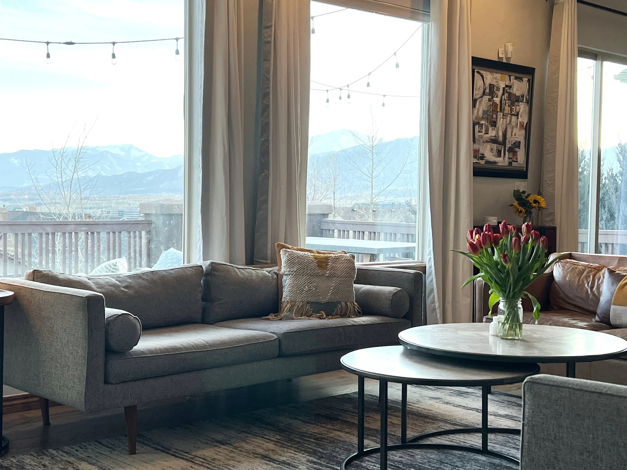 A living room with a gray sofa, a half-moon-shaped coffee table with a vase of pink tulips, and a large window with a mountain view.
