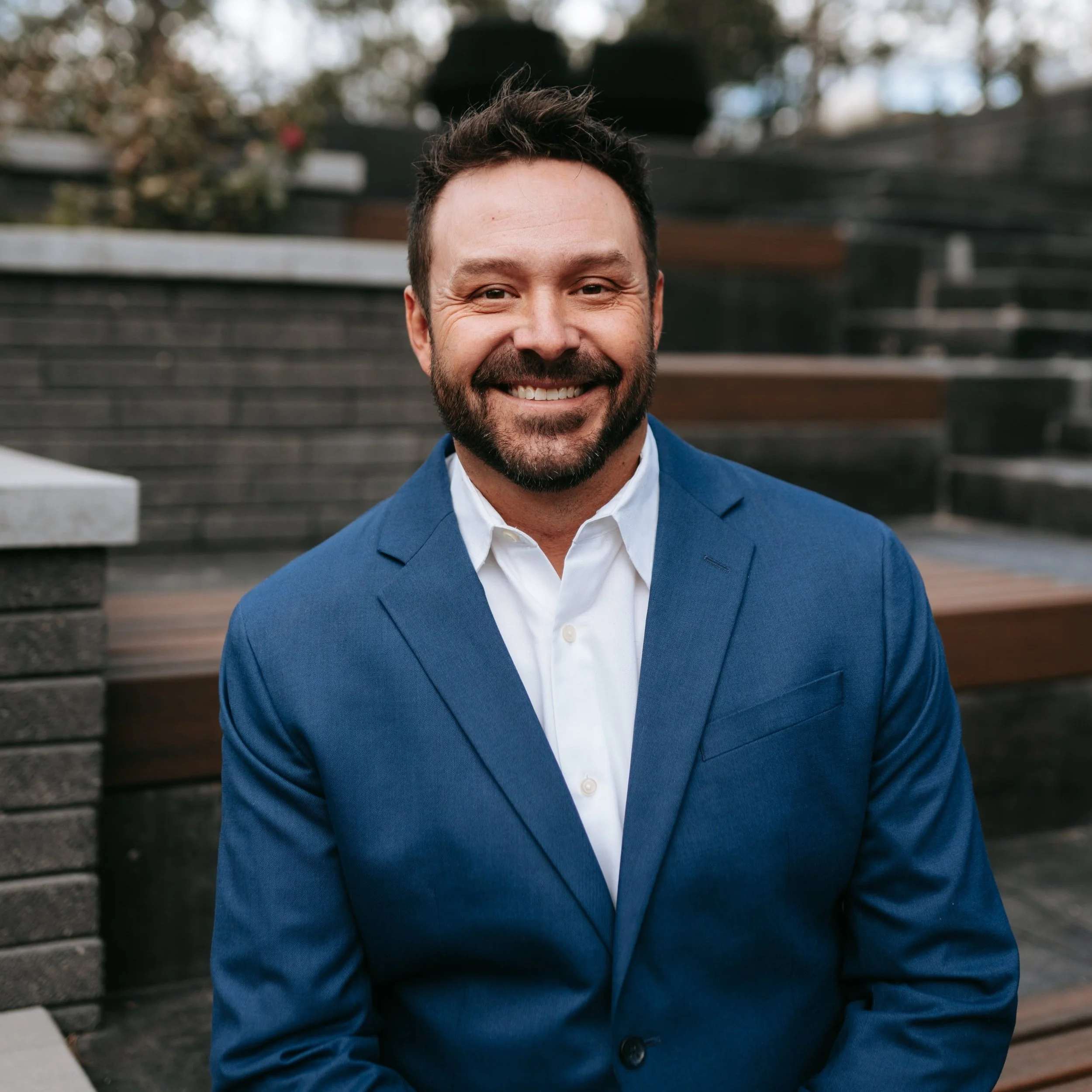 A smiling man with dark hair and a beard, wearing a blue suit jacket and a white dress shirt, standing outdoors on steps with blurred background of trees and steps.