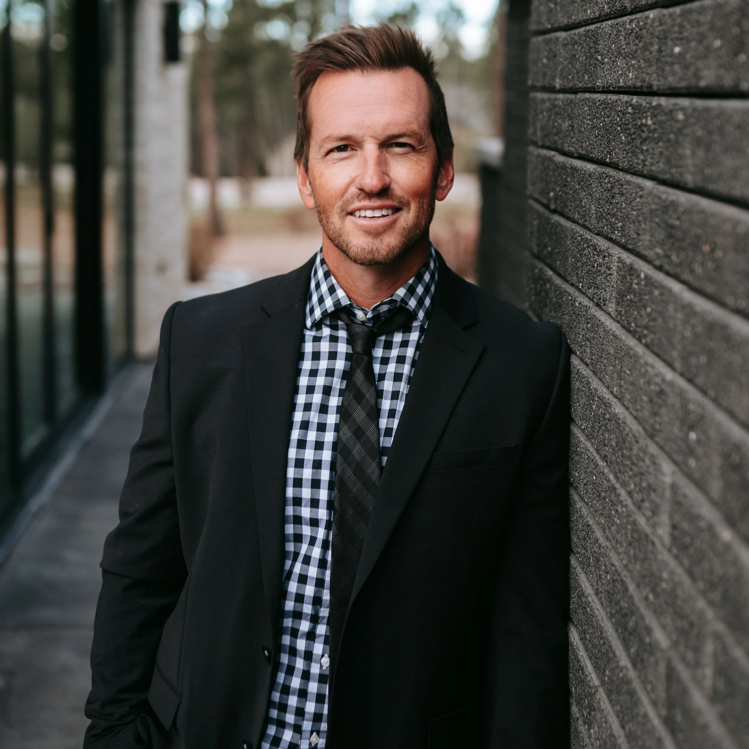 A man in a black suit and checkered shirt smiling while leaning against a wooden wall outdoors.