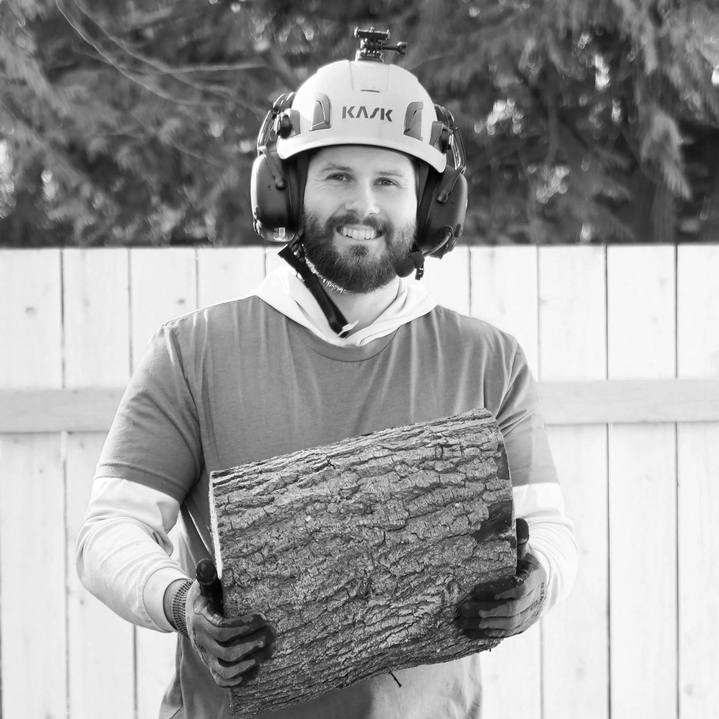 A man with a beard wearing a helmet, safety gear, and gloves, holding a large piece of wood, standing outdoors in front of a fence and trees.