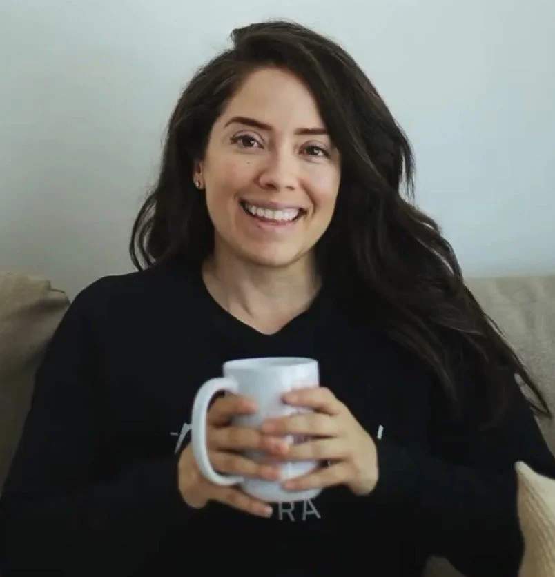 A woman with long dark hair smiling, holding a white mug with both hands, sitting on a beige couch in a light-colored room.