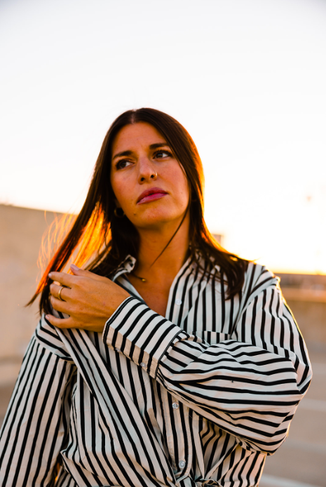 A woman with long dark hair wearing a black and white striped shirt, standing outdoors during sunset, touching her shoulder and looking thoughtful.