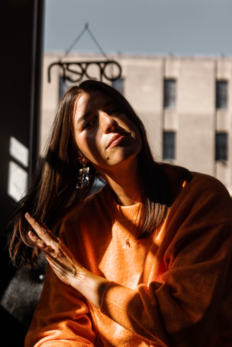 Young woman with brown hair in sunlight, wearing an orange sweater, near window with building outside.