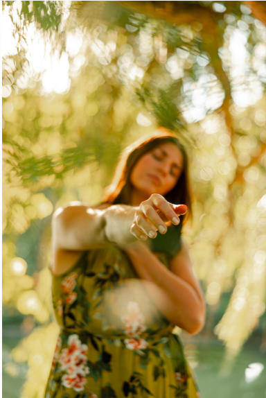 Woman in a floral dress reaching out with her hand in an outdoor setting with trees and sunlight.