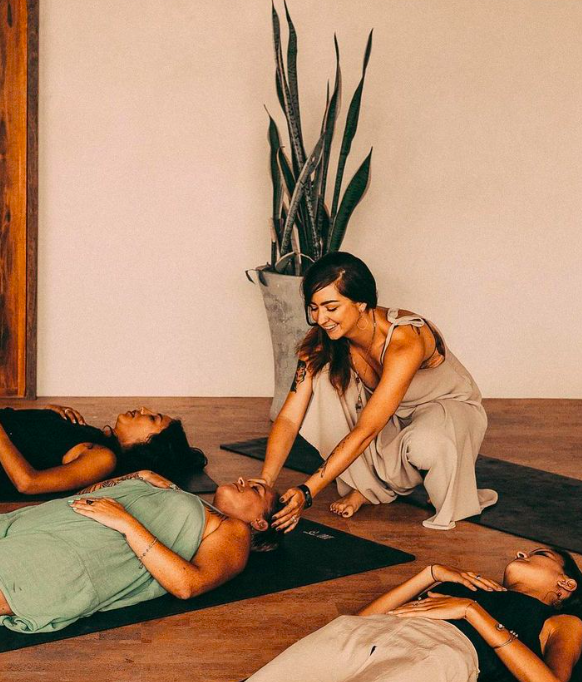Women practicing yoga in a wellness studio with a yoga mat, a large plant, and a plain wall.