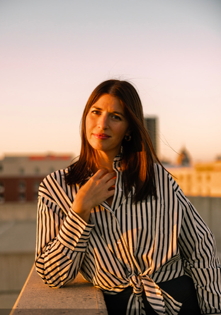 Woman with striped shirt standing outdoors during sunset, with city buildings in the background.